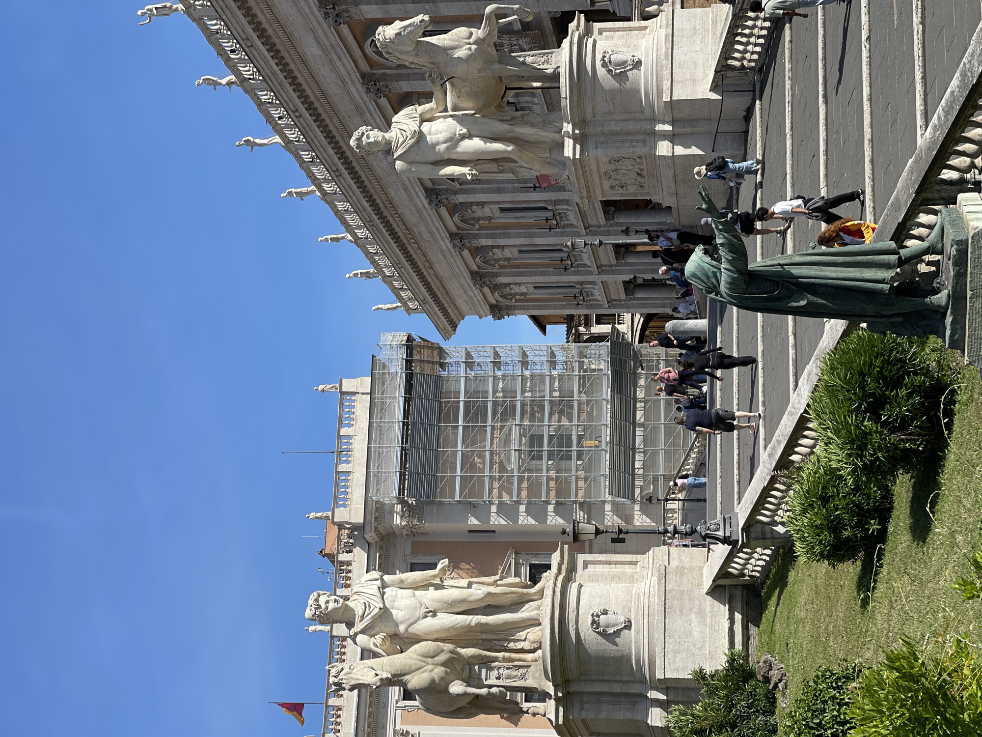 The image shows the Piazza del Campidoglio in Rome, Italy. Two statues of Castor and Pollux stand at the top of the Cordonata stairs, flanking the Palazzo Senatorio. Scaffolding covers part of the facade of the Palazzo Senatorio. A bronze statue of Emperor Marcus Aurelius is to the right of the Cordonata stairs. People are walking up and down the stairs. Greenery grows along the sides of the staircase.
