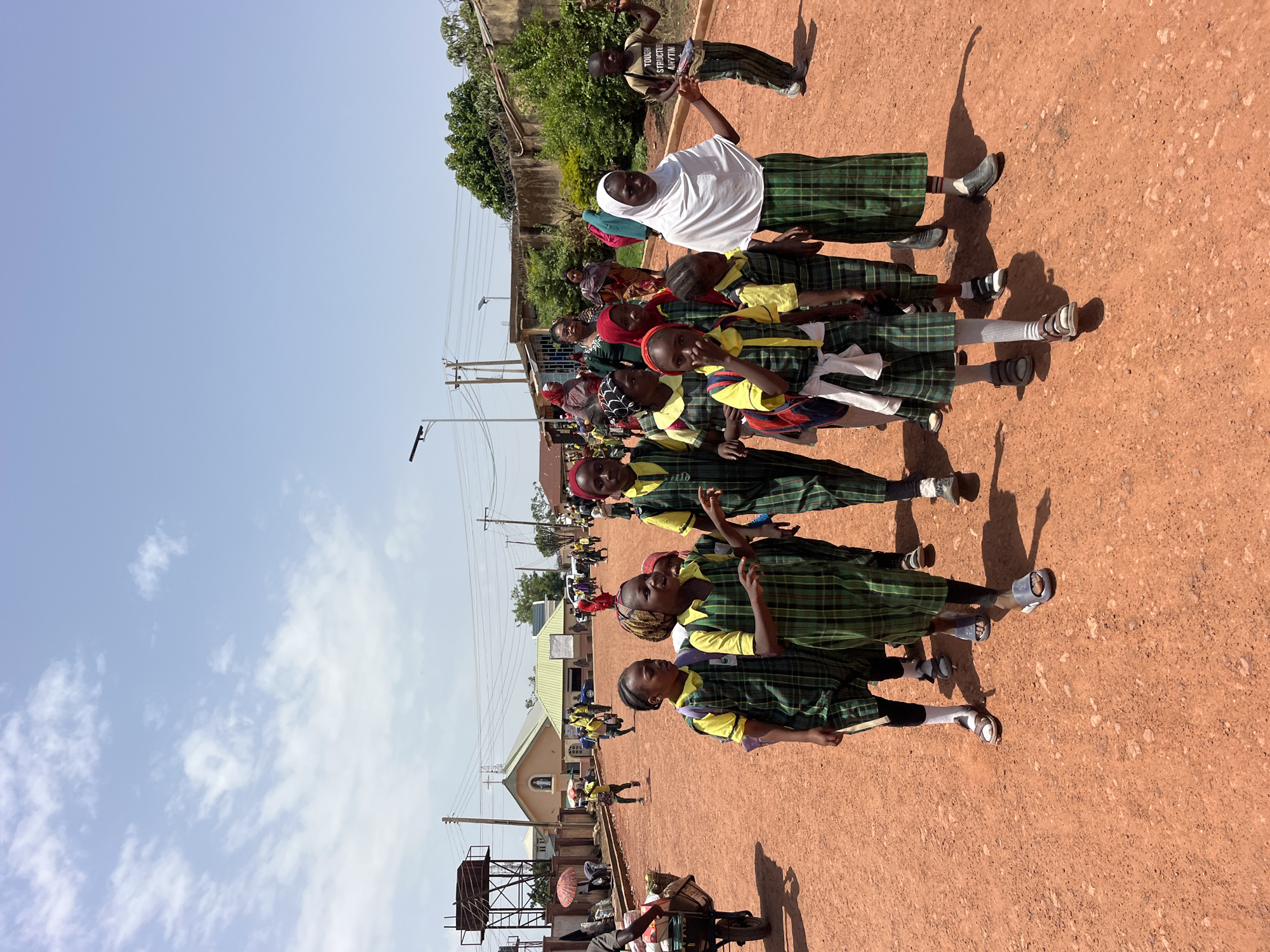 A group of children in plaid school uniforms are walking on a dirt road. The children are of various ages and are walking in the same direction. Some of the children have backpacks on. In the background, there are buildings, trees, and a church. The sky is blue with some clouds. The overall scene appears to be a sunny day in an outdoor setting.