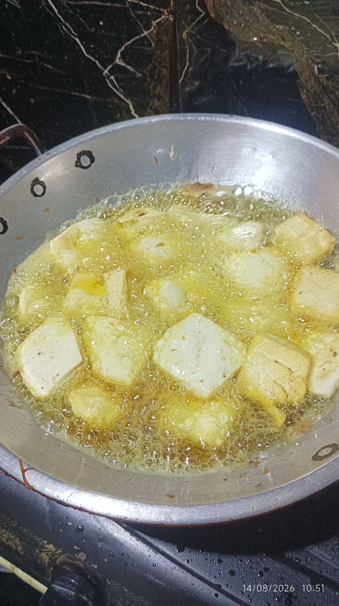A close-up, overhead shot captures cubes of tofu being deep-fried in a wok. The golden-brown cubes bob in bubbling hot oil, some showing a slightly lighter shade indicating they are further along in the frying process. The wok itself is made of metal and shows signs of use, with some dark specks and wear marks on its surface. The background is a dark, marbled surface, possibly a countertop. The scene appears to be in a kitchen in Tasikmalaya, Indonesia. The date and time stamp "14/08/2026 10:51" is visible in the lower right corner, suggesting the image was taken on August 14, 2026, at 10:51 AM.