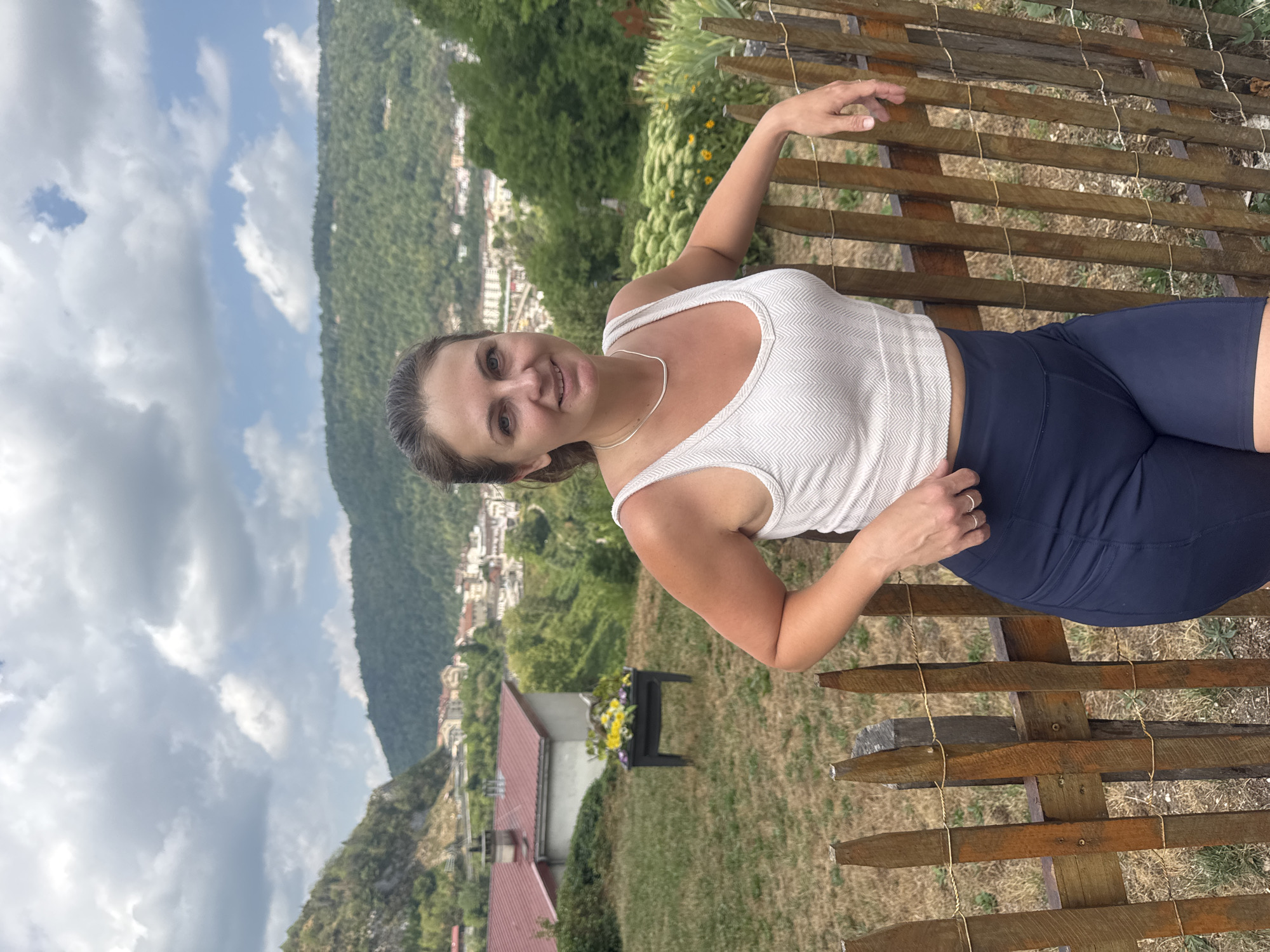 A woman stands outdoors leaning against a wooden fence. She is wearing a white tank top and dark blue shorts. Her hair is pulled back in a ponytail. Behind her, a town is visible nestled in a valley, with buildings and trees stretching up the hillsides. The sky is cloudy. The area appears to be a residential neighborhood with houses and gardens. The woman is the central focus of the image.
