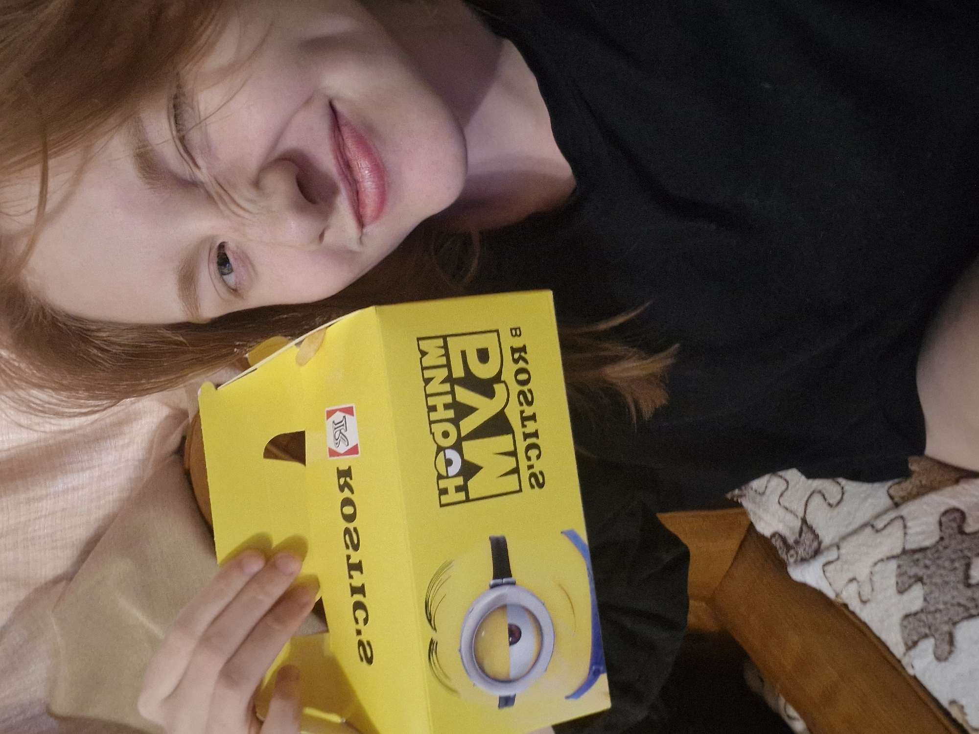 The image shows a young woman with red hair and fair skin, holding a yellow box featuring a Minion character. She is winking and smiling, with dimples visible on her cheeks. The box has text that appears to be in Russian and is partially obscured. The woman is wearing a black shirt. The background is blurred, showing what looks like a light-colored blanket or quilt with a puzzle piece pattern, and possibly wooden furniture. The scene appears to be indoors. There is no indication of the location in Yekaterinburg, Russia.