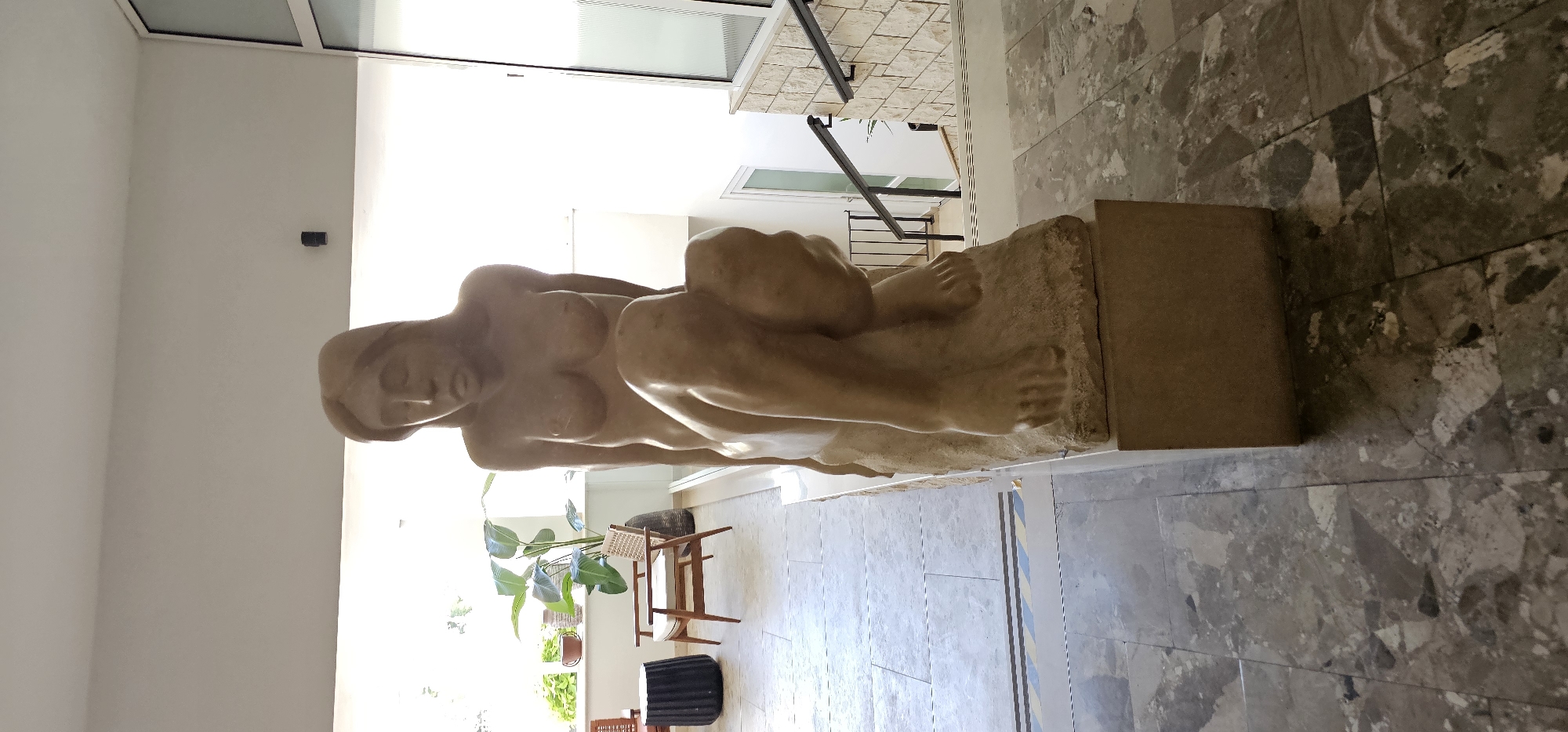 A nude, light-brown sculpture of a woman is displayed on a stone pedestal in the city of Grad Supetar, Croatia. The sculpture depicts a woman with her head bowed, her legs drawn up to her chest, and her arms wrapped around her knees. Her body is lean and muscular, with subtle details in the musculature of her legs and torso. The sculpture's surface appears smooth, suggesting it's made of polished stone or a similar material.
The sculpture is positioned on a low, rectangular pedestal in what appears to be an indoor or semi-outdoor public space. The background reveals a bright, airy environment with natural light streaming in from large windows. To the left of the sculpture, there are two chairs and a small dark cylindrical object, possibly a stool or side table, on a tiled floor. Further back, through the windows, lush green plants are visible, suggesting a garden or an atrium.
To the right of the sculpture, a staircase with black metal railings descends. The walls surrounding the staircase are clad in light-colored stone, providing a textured contrast to the smoother surfaces elsewhere. The floor in the foreground and around the pedestal is made of large, multi-colored stone tiles with a polished finish, reflecting the ambient light. The overall impression is one of a modern, minimalist setting designed to showcase the artwork.