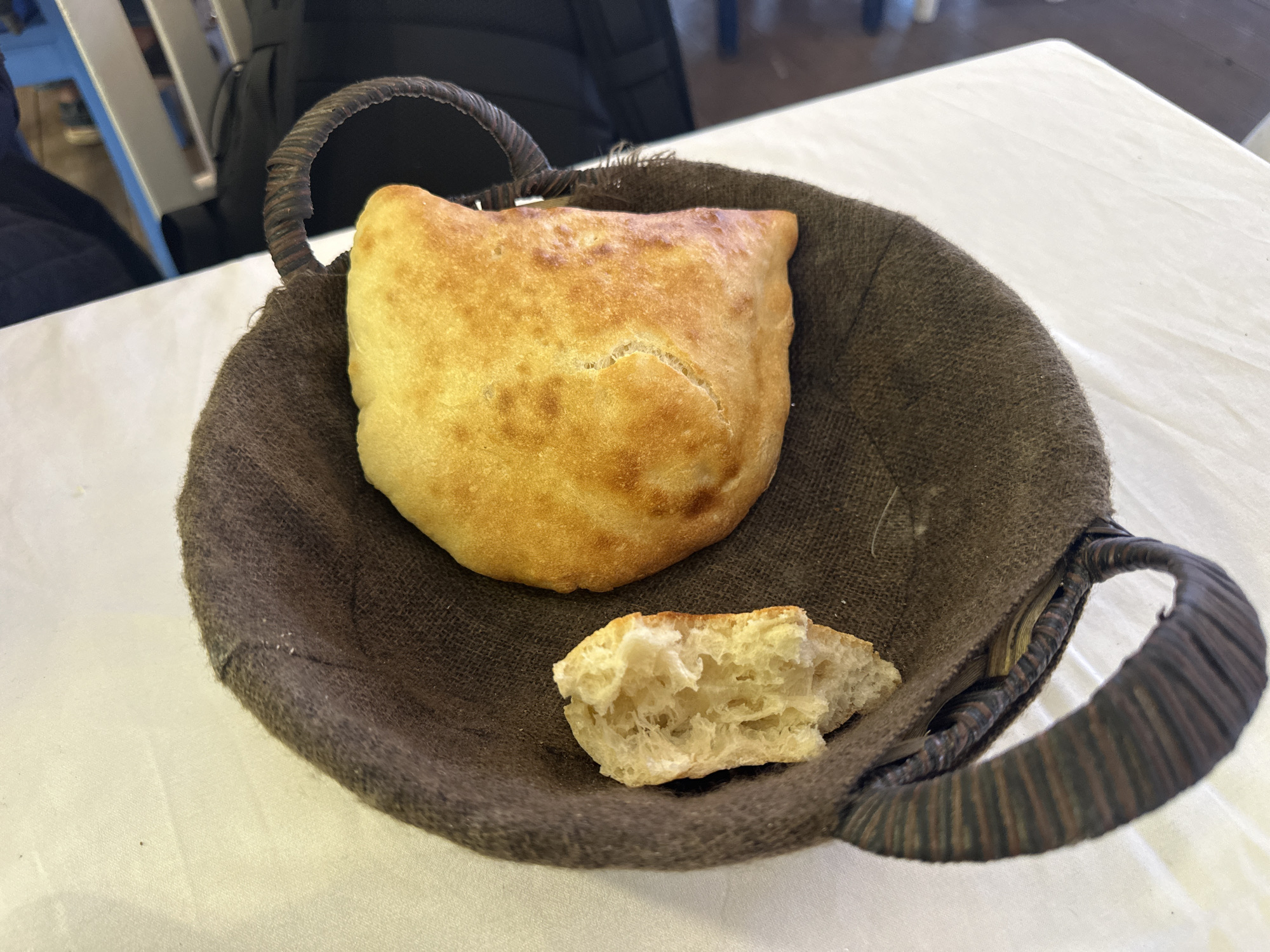 The image shows a basket of bread on a table. The basket is made of a woven material and lined with brown fabric. Inside the basket, there is a whole piece of golden-brown bread, along with a smaller, torn piece, revealing its soft, airy interior. The table is covered with a white tablecloth. The image was taken in the city of Fethiye, Turkey.