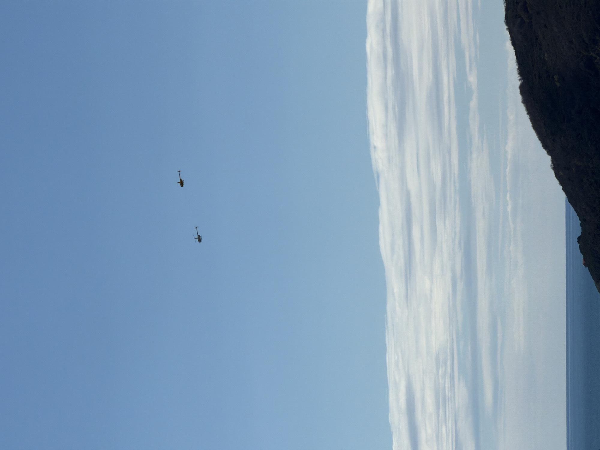 The image shows two yellow helicopters in a clear blue sky near Budva, Montenegro. A long, thin cloud formation stretches vertically along the right side of the frame, adjacent to a dark, rocky landmass. A hint of the Adriatic Sea is visible at the base of the cloud formation.