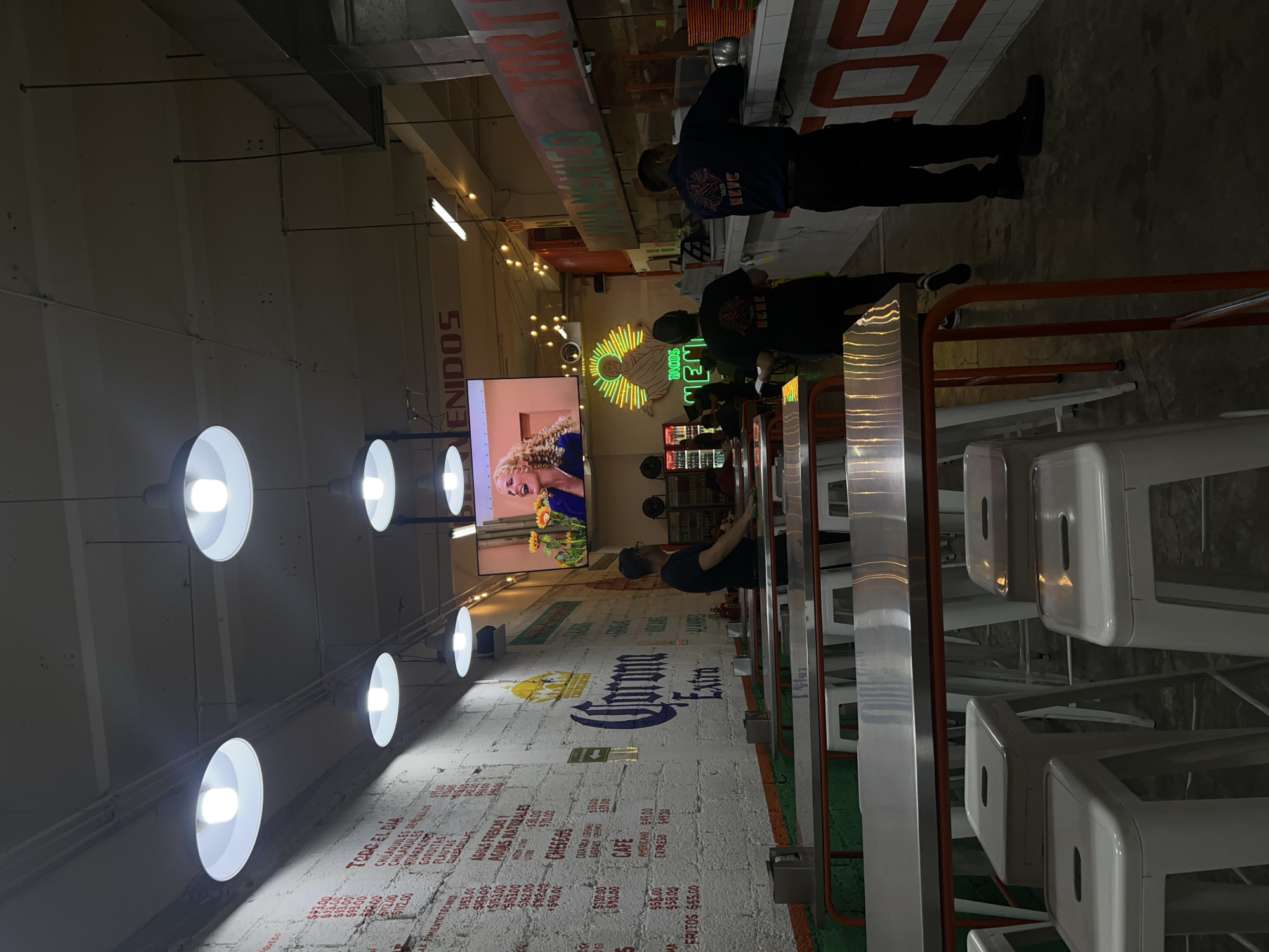 The image shows the interior of a restaurant in Mexico City, Mexico. The restaurant has a casual, industrial feel with exposed ductwork and brick walls. There are people lined up at a counter, presumably ordering food. The menu is written on the wall in Spanish. In the background, there is a large television screen displaying an image, and a neon sign with the words "TACOS MEM". The seating area has rows of tables and white stools. The lighting is provided by industrial-style pendant lights. The overall atmosphere is lively and informal.