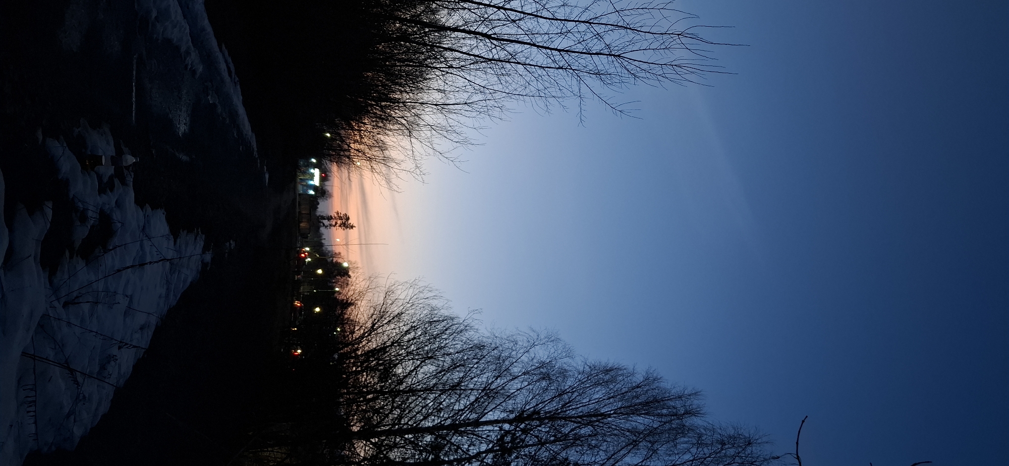 The image, taken in Chaikovsky, Russia, shows an evening scene with a predominantly blue sky transitioning to a pinkish hue near the horizon. Silhouetted bare trees frame the top and bottom edges of the image. On the left, a snow-covered embankment or hillside is visible, leading to a dimly lit area with what appears to be buildings and illuminated streetlights. The location seems to be on the outskirts of the city, as indicated by the natural elements and the presence of urban lighting in the distance. No people are visible.