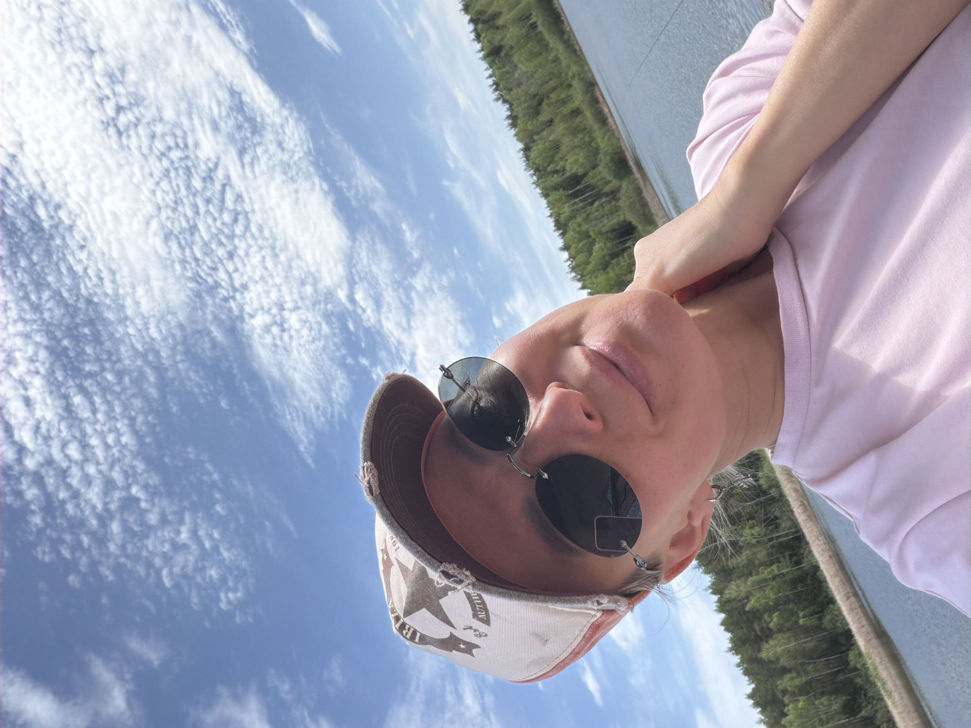 A woman wearing sunglasses and a baseball cap is positioned with her head tilted, likely near a body of water lined with trees, possibly in Chaikovsky, Russia. She is wearing a light-colored top and her hand is touching her neck. The sky has a clear blue with scattered clouds.