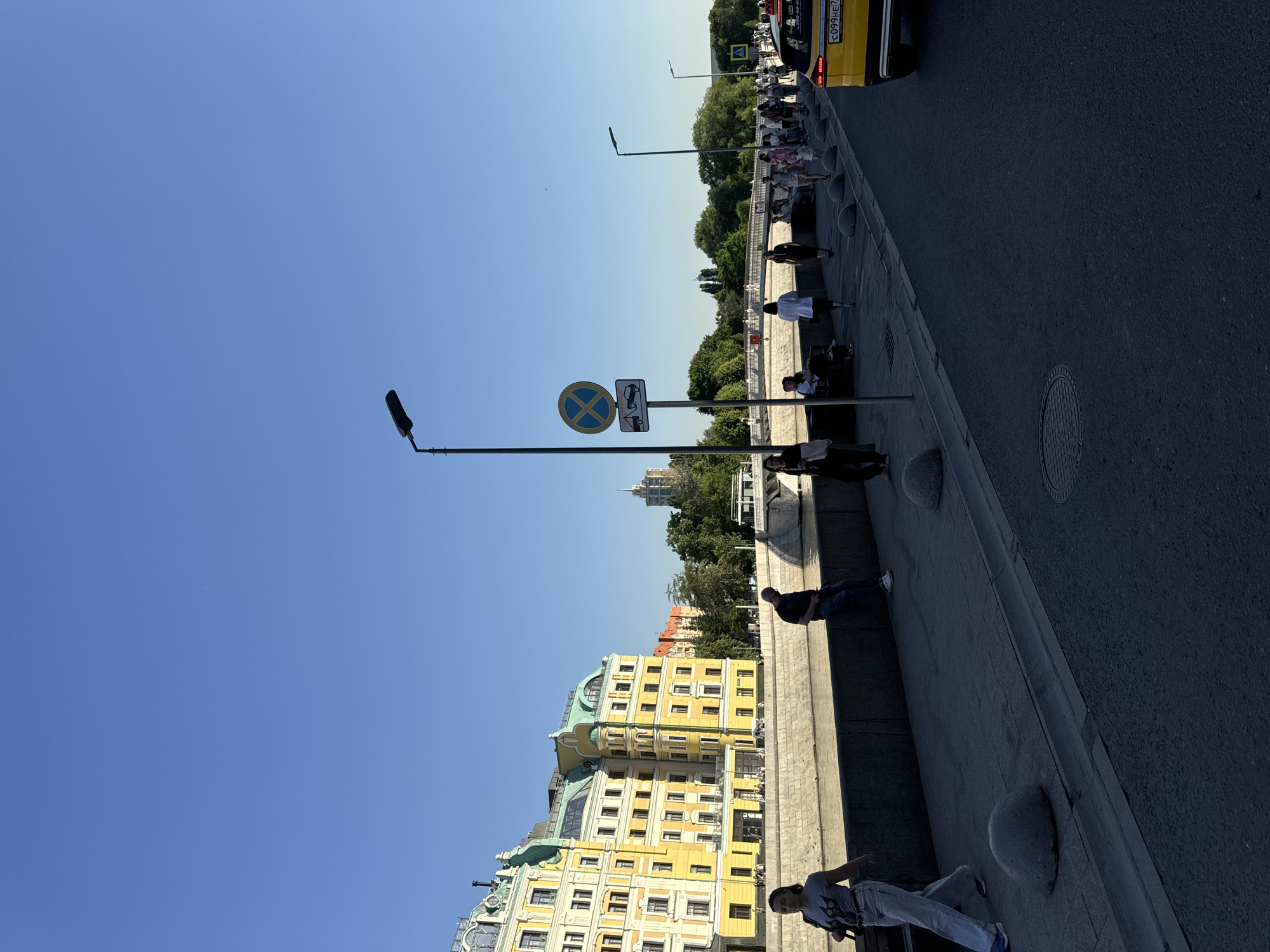 This image captures a clear, sunny daytime scene along the Moskva River in Moscow, Russia. The perspective is from an embankment, looking across the river towards the Hotel Baltschug Kempinski Moscow. In the upper portion of the image, the sky is a uniform, clear blue with no visible clouds. The prominent architectural feature in the background on the left is the Hotel Baltschug Kempinski Moscow, a large, ornate building with yellow facades, multiple windows, and distinctive green, domed roofs with white decorative elements. To its right, other buildings with reddish-orange tones are partially obscured by dense green trees. The Moskva River, with its murky, light brown water, flows through the mid-ground, separating the foreground embankment from the background buildings. A dark grey concrete wall forms the immediate river embankment. In the foreground and mid-ground, a paved pedestrian sidewalk, constructed from grey rectangular tiles, runs parallel to a dark asphalt road on the right. Several spherical, grey concrete bollards are spaced along the edge of the sidewalk, separating it from the road. A circular manhole cover is visible on the asphalt road in the lower right. A tall, dark grey street lamp with a horizontal light fixture stands in the mid-ground, slightly to the right of the center. Attached to this pole are two traffic signs: a circular "No Stopping" sign (blue background with a red diagonal cross) and a supplementary rectangular sign depicting a tow truck towing a car, indicating a "No Stopping, Tow-Away Zone." Further down the road to the right, another identical street lamp is visible, along with a yellow pedestrian crossing sign partially seen. Numerous individuals populate the scene. In the foreground left, a woman with dark hair, wearing a grey graphic t-shirt, light blue jeans, and blue sneakers, walks away from the camera, carrying a dark handbag. In the mid-ground, a man in a dark t-shirt and jeans is seated on the concrete embankment wall, looking towards the river. Further along the embankment and across a white bridge in the distance, a continuous stream of people can be observed walking and congregating. A portion of a yellow car, identified as a taxi by its color and a visible Russian license plate reading "c099HE 77" (where "77" denotes the Moscow region), is parked or driving on the road in the far right foreground, facing away from the viewer. In the far background, a white boat or ferry