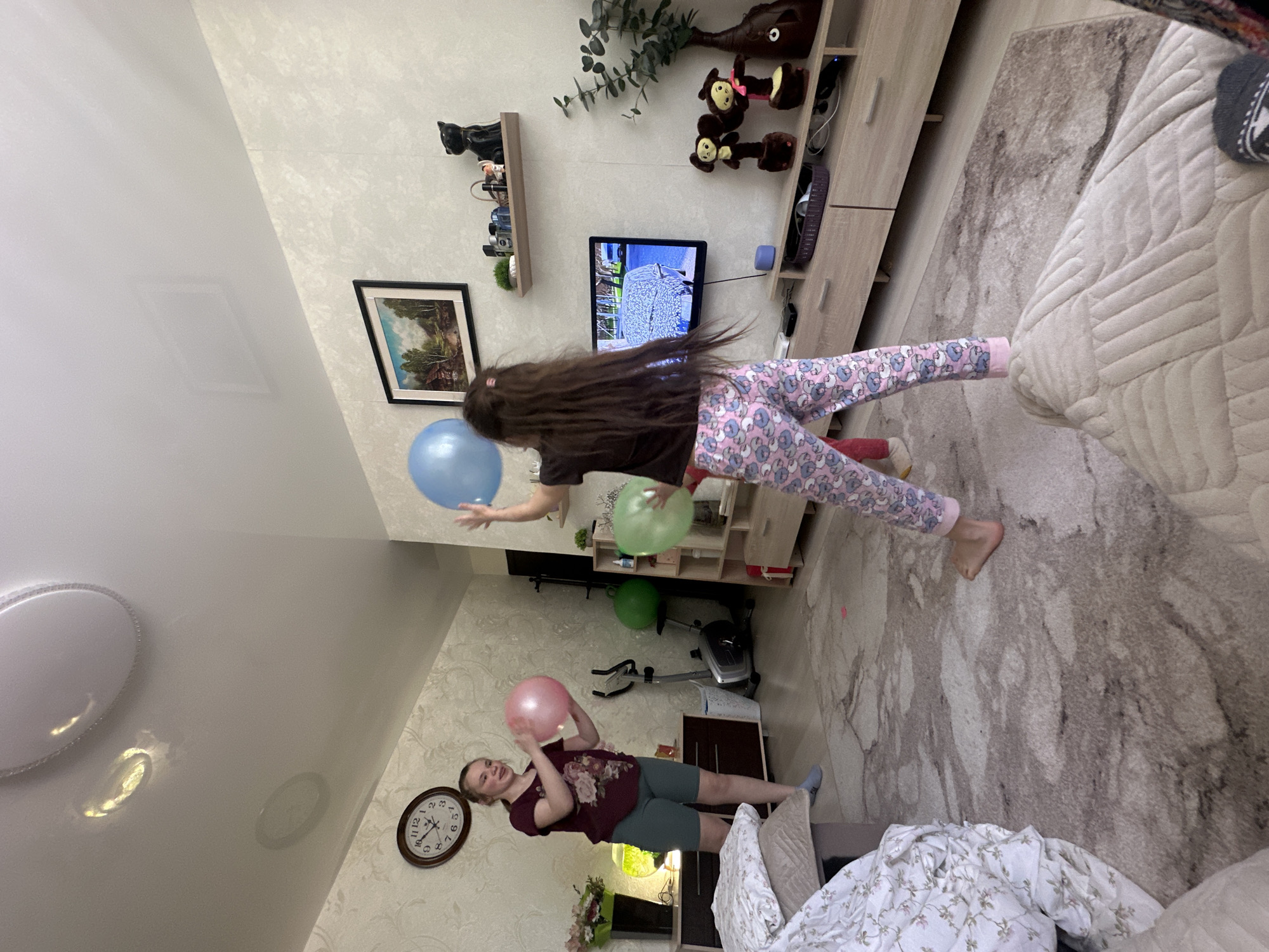 The image depicts an interior scene, likely a residential space, in what appears to be the city of Uva, Russia. A girl, with long brown hair, dressed in a dark shirt and patterned pants, is airborne. She is positioned near a television and a set of shelves containing decorative objects. She is playing with a blue and a green balloon. Below her, another person, possibly a woman, wearing a maroon t-shirt and green shorts, is lying on a sofa and playing with a pink balloon. There is a clock on the wall, showing the time. The room contains typical domestic furnishings, including a patterned rug, a sofa, and some home decor. The environment is a casual, lived-in setting.