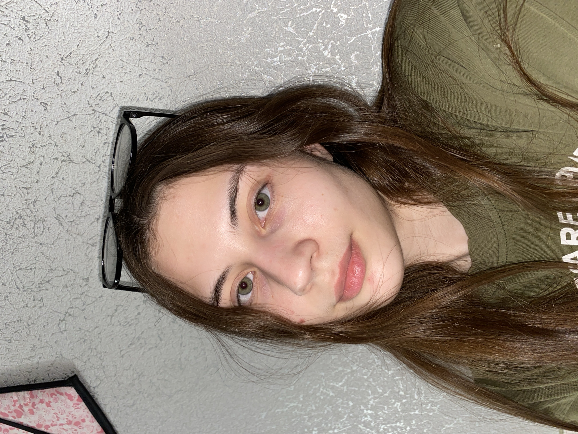 A young woman with long brown hair and light olive green eyes lies on her side against a textured grey wall. She wears a green t-shirt, and a pair of black-rimmed glasses rests on the wall beside her head. The bottom corner of a patterned pink and black object is visible in the lower left of the frame. The location of the image is not discernible from the provided details, and there's no confirmation it is in Votkinsk, Russia.