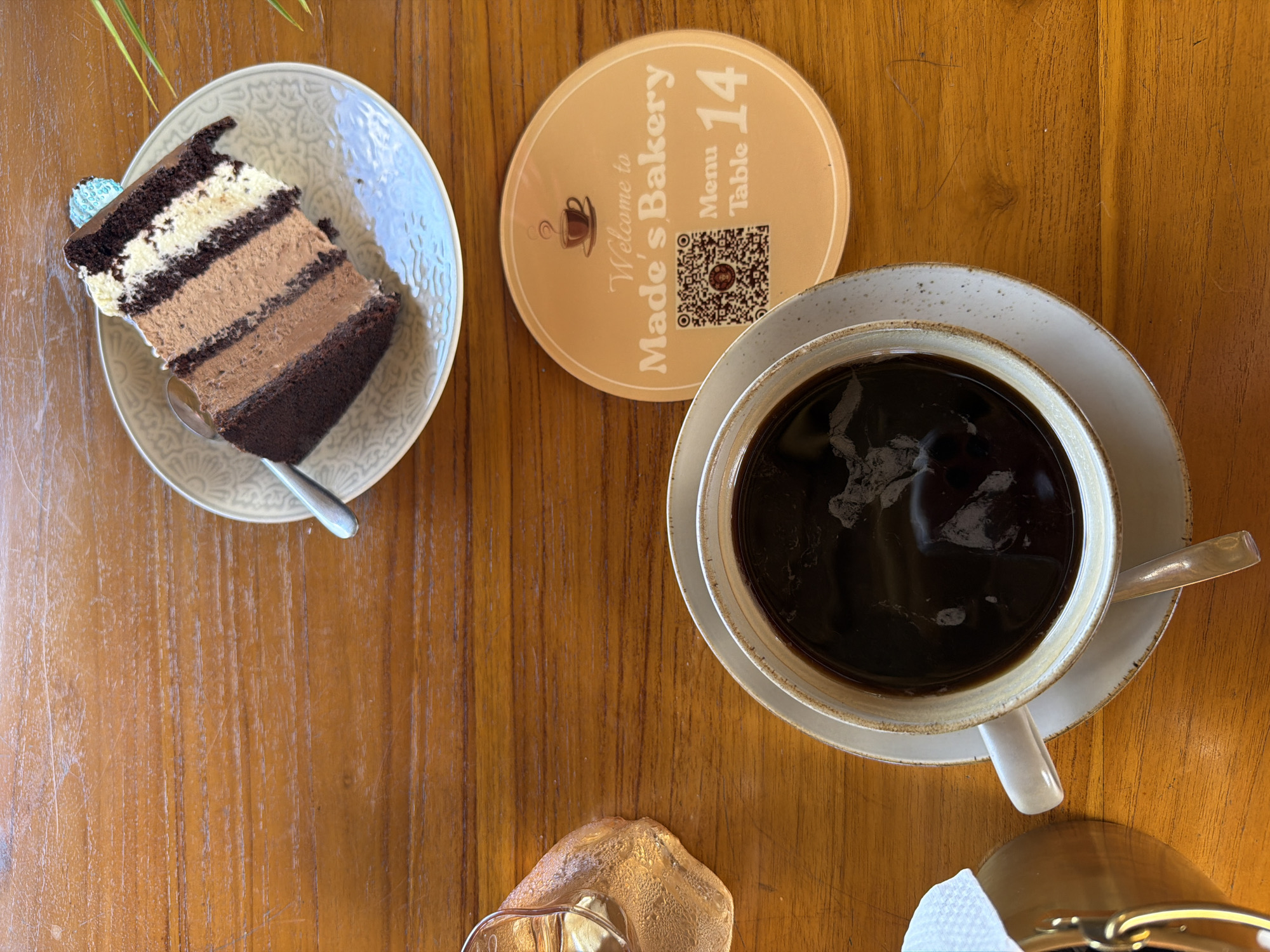 The image captures a tabletop scene in what appears to be a cafe or bakery.
**Objects:**
* A slice of layered chocolate cake on a light-colored, patterned plate with a spoon.
* A round coaster with the inscription "Welcome to Made's Bakery Menu Table 14" and a QR code.
* A cup of black coffee on a saucer.
* A small pitcher (partially visible) likely containing cream or syrup, placed at the bottom left corner of the shot.
* Napkins in a gold colored holder in the lower right corner.
**Environment:**
* The objects are placed on a wooden table with a polished surface, potentially made of teak.
* A green plant is partially visible in the upper left corner.
**Location and Context:**
* The table coaster indicates the location is "Made's Bakery". Based on the prompt, the city is Kuta Selatan, Indonesia. The scene is a typical cafe setting with cake and coffee.