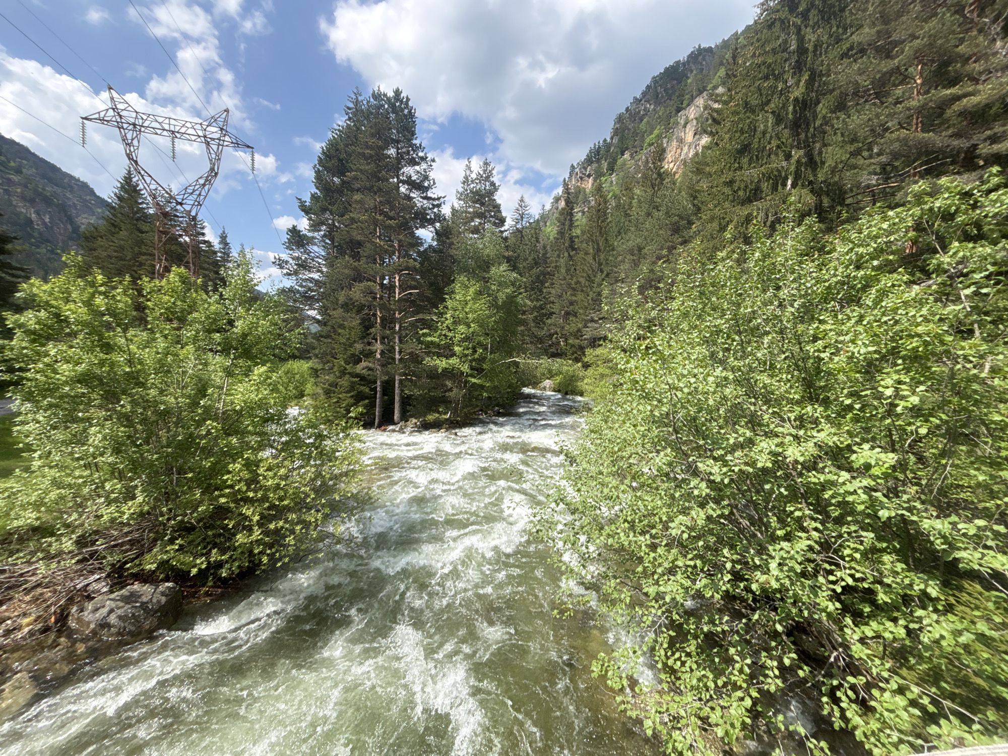 The image displays a rugged natural landscape in Beli Iskar, Bulgaria, featuring a fast-flowing mountain river under a partially cloudy sky. A turbulent river with significant white water and a greenish-brown hue occupies the central and lower portions of the frame, moving from the upper right towards the foreground. Lush, vibrant green deciduous bushes and trees line both riverbanks, some branches overhanging the water.
On the left side, a large, rust-colored, lattice-structured electrical transmission tower is prominently positioned against the sky, supporting multiple power lines that stretch horizontally. Beyond the immediate river course, dense forests comprised primarily of conifer trees, along with some deciduous species, cover the ascending terrain. Steep, tree-clad mountains frame the river valley in the background, with the mountain on the right exhibiting areas of visible rock face among the foliage. The sky is blue with scattered, white cumulus clouds. No people or animals are present in the scene.