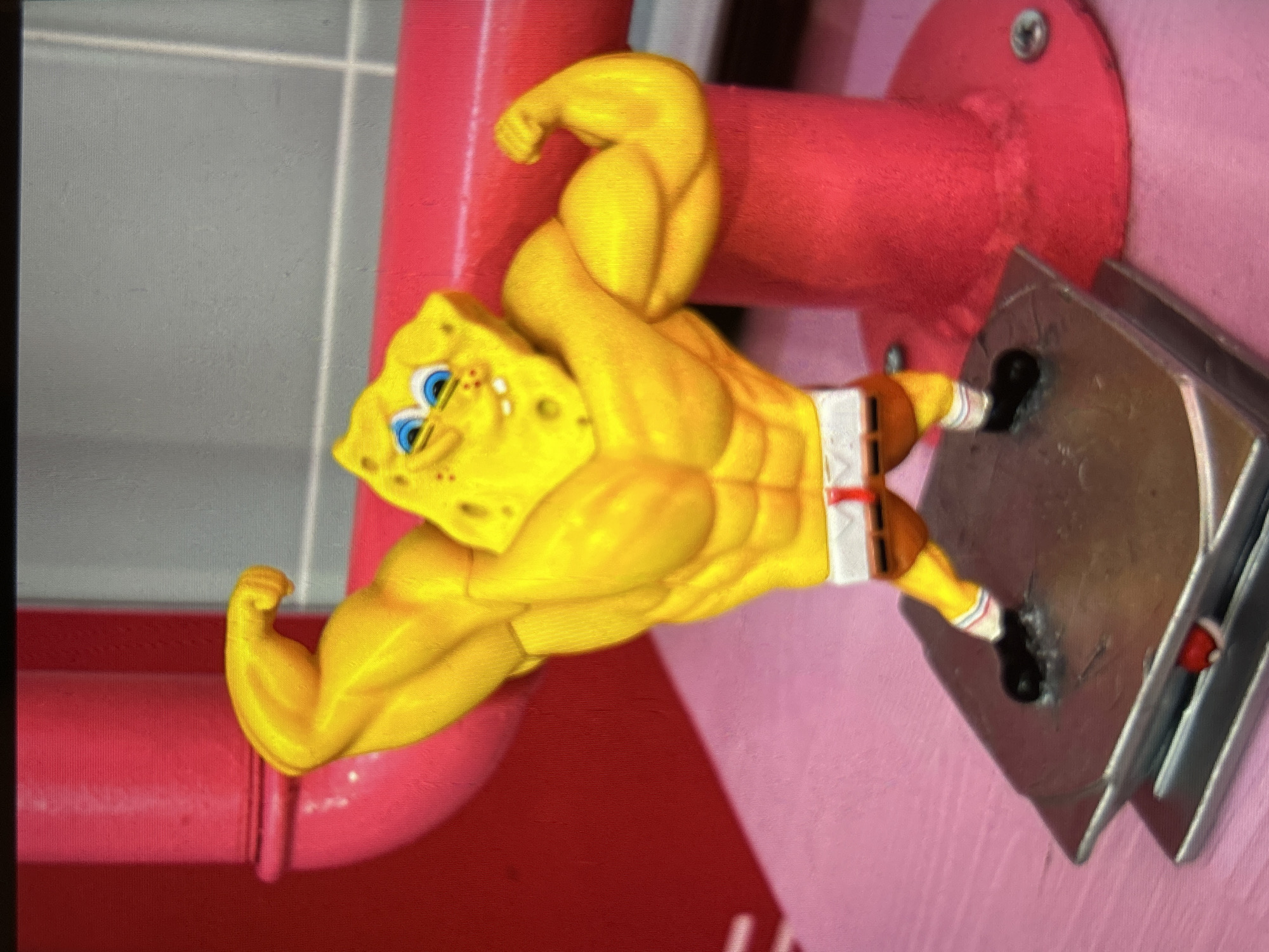 A muscular figurine of SpongeBob SquarePants stands flexing his biceps. He has a yellow, porous body, blue eyes, and a wide grin. He is wearing brown shorts with a white waistband and red trim, and black shoes with white and red striped socks. The figurine is standing on a metallic base. In the background, there are large, bright pink cylindrical objects, possibly plumbing or decorative elements, and a pink, textured surface beneath the figurine. The overall environment appears to be a playful or display setting, possibly a store or a themed room, with a slightly out-of-focus background suggesting a shallow depth of field.