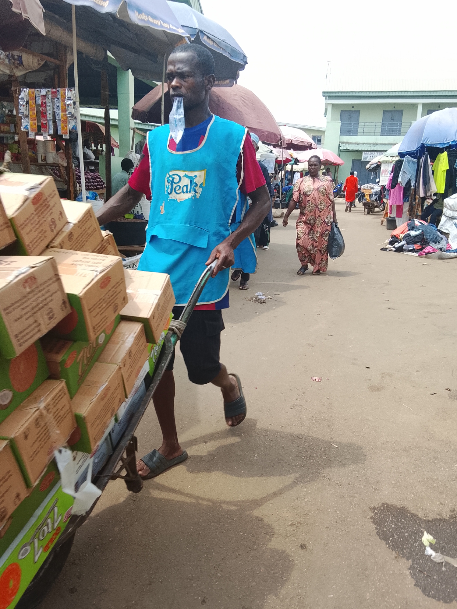 This image captures a vibrant and busy market scene in Akwanga, Nigeria, under a bright, overcast sky.
In the foreground, a dark-skinned man is the primary subject, actively pushing a metal handcart loaded high with numerous cardboard boxes. He wears a red t-shirt beneath a light blue apron, prominently featuring the "Peak" brand logo in white. A clear plastic bag or bottle top is held firmly in his mouth. He is dressed in dark shorts and flip-flops, with his legs showing motion as he moves the heavy cart. The boxes on the cart are light brown, with some featuring green labels that clearly display "Tio 7" and what appears to be a coconut illustration, suggesting tropical produce or products.
In the mid-ground, a dark-skinned woman walks towards the viewer, carrying a black plastic bag in her left hand. She is dressed in a patterned, loose-fitting, reddish-brown dress. Beyond her, the market extends with various stalls, shaded by large, colorful umbrellas. Clothing items are visible hanging for sale.
The background reveals a multi-story building with a light green facade and several windows, bearing a sign that reads "MADU TEX". Another stall in the distance has a visible "POS" sign. The ground is unpaved, appearing to be packed earth or dirt, showing some signs of wear and possibly wet patches. The overall atmosphere is one of commerce and movement, typical of an outdoor market.