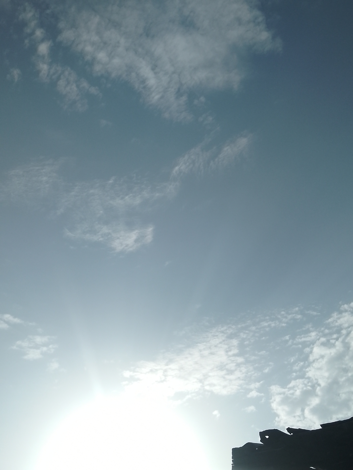 The image shows a silhouetted dark structure against a bright sky. The structure appears to be rocky or made of dark, uneven material, possibly a building or natural formation, occupying the lower right portion of the frame. Above this structure, the sky is a gradient of light blue, with scattered white clouds. A bright light source, likely the sun, is visible in the lower left quadrant, creating a strong glare and washing out details in that area. The overall scene is natural, with no visible people or specific urban landmarks to indicate a precise city location.