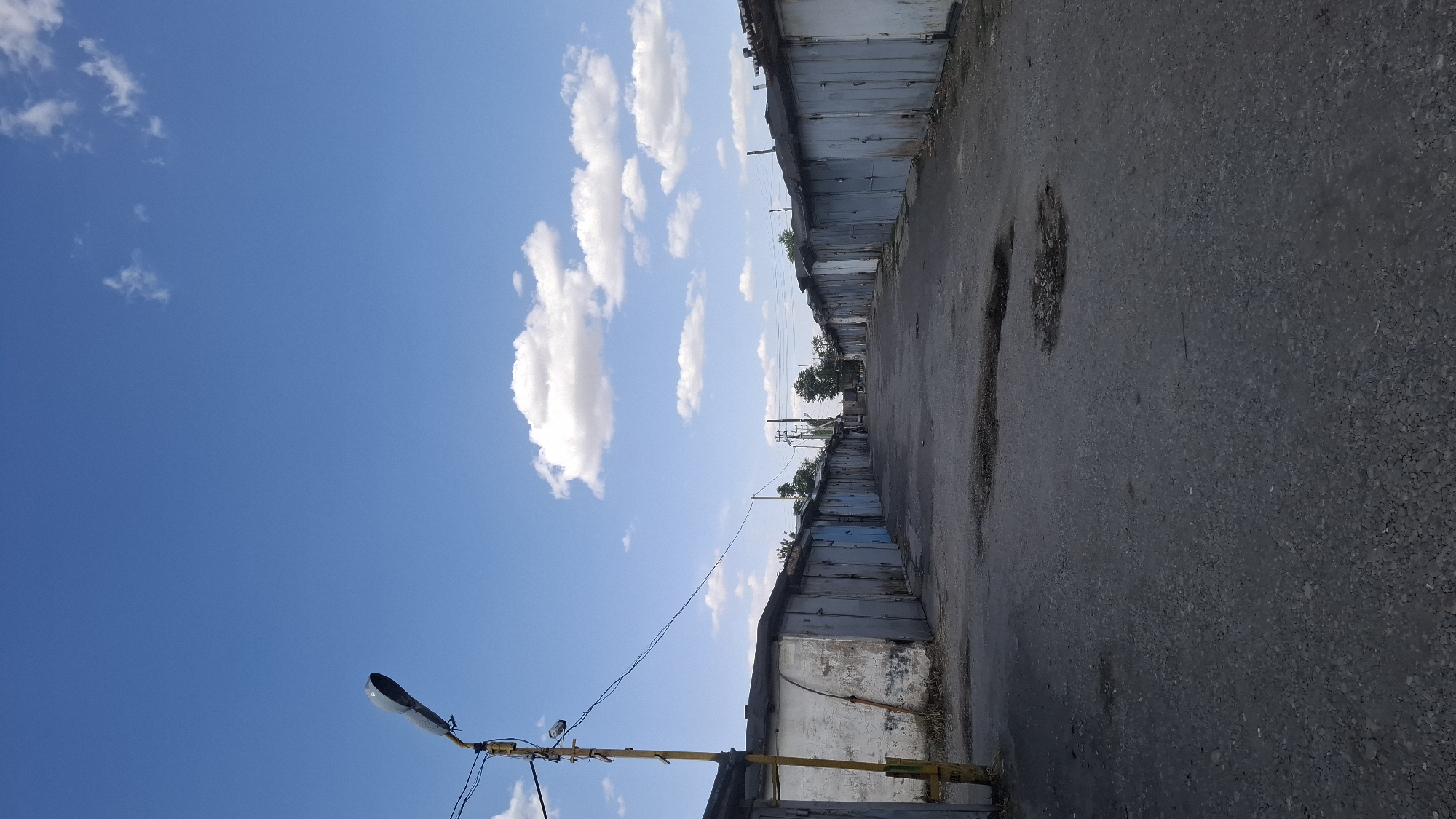 The image depicts a row of weathered garages under a bright blue sky with scattered white clouds. The garages are made of concrete and metal, with some showing signs of age and disrepair. The ground in front of the garages is a dark asphalt or gravel surface with several shallow puddles. A street lamp with a visible bulb is positioned on the left side of the image, attached to a yellow pole. The scene suggests an industrial or storage area, possibly a garage cooperative or a similar facility, located in an urban environment. The overall impression is one of utilitarian simplicity and everyday life.