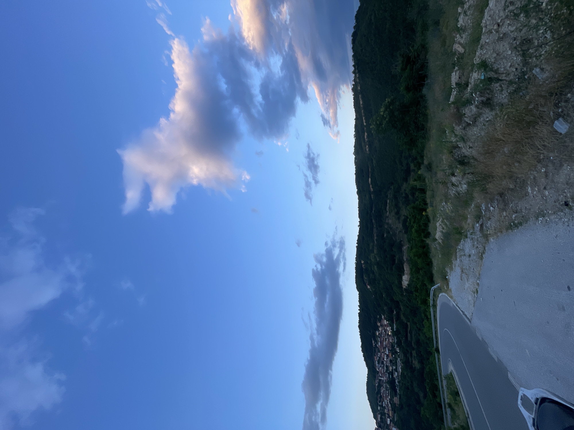 The image depicts a twilight view of a winding road and a hillside town. In the foreground, a paved road curves to the left, with a worn, gravelly shoulder on the right. The road appears to be a two-lane street with a white dividing line. Beyond the road, a densely wooded hillside rises, dotted with buildings that form a town. The houses are predominantly red-roofed and clustered together. The sky is a deep blue, with wispy white and gray clouds scattered across it. The overall impression is of a serene, rural landscape at dusk. The location is identified as Bogolin, Bulgaria. There are no people or animals visible in the image.
