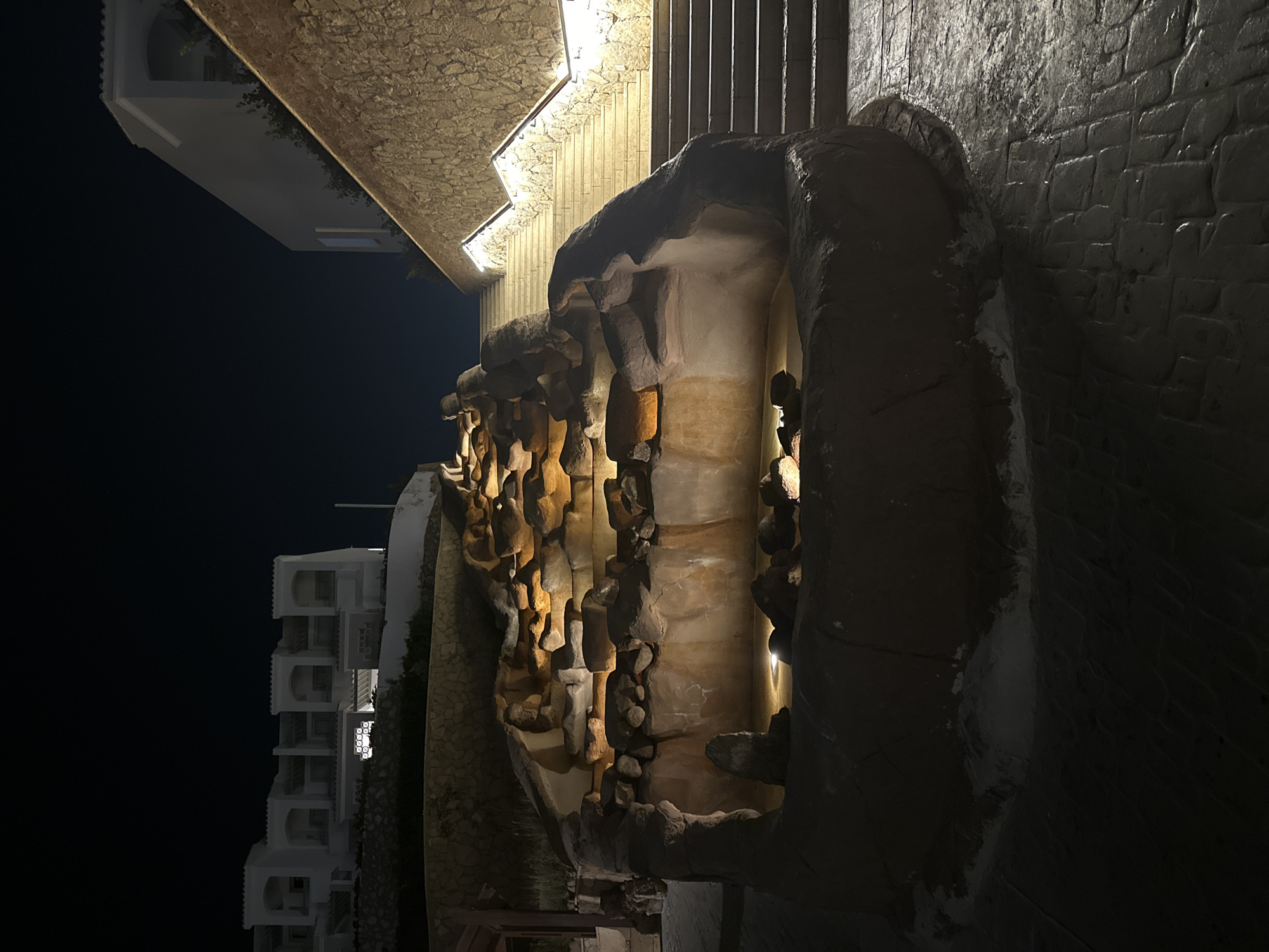 This night-time image captures an outdoor architectural and landscape scene in Unknown, Unknown. A multi-tiered artificial rock waterfall or fountain, illuminated with warm yellow lights embedded within its structure and reflecting on pooled water, occupies the central midground. To the left, a multi-story white building featuring a series of arched windows and balconies is situated on a terraced hillside, supported by a stone retaining wall. Several windows in the building display interior illumination. To the right, a wide, upward-sloping staircase is visible, brightly lit by linear fixtures integrated into its handrails, running alongside a textured stone wall. The ground in the immediate foreground is paved with dark, patterned stones. No individuals are present in the scene.