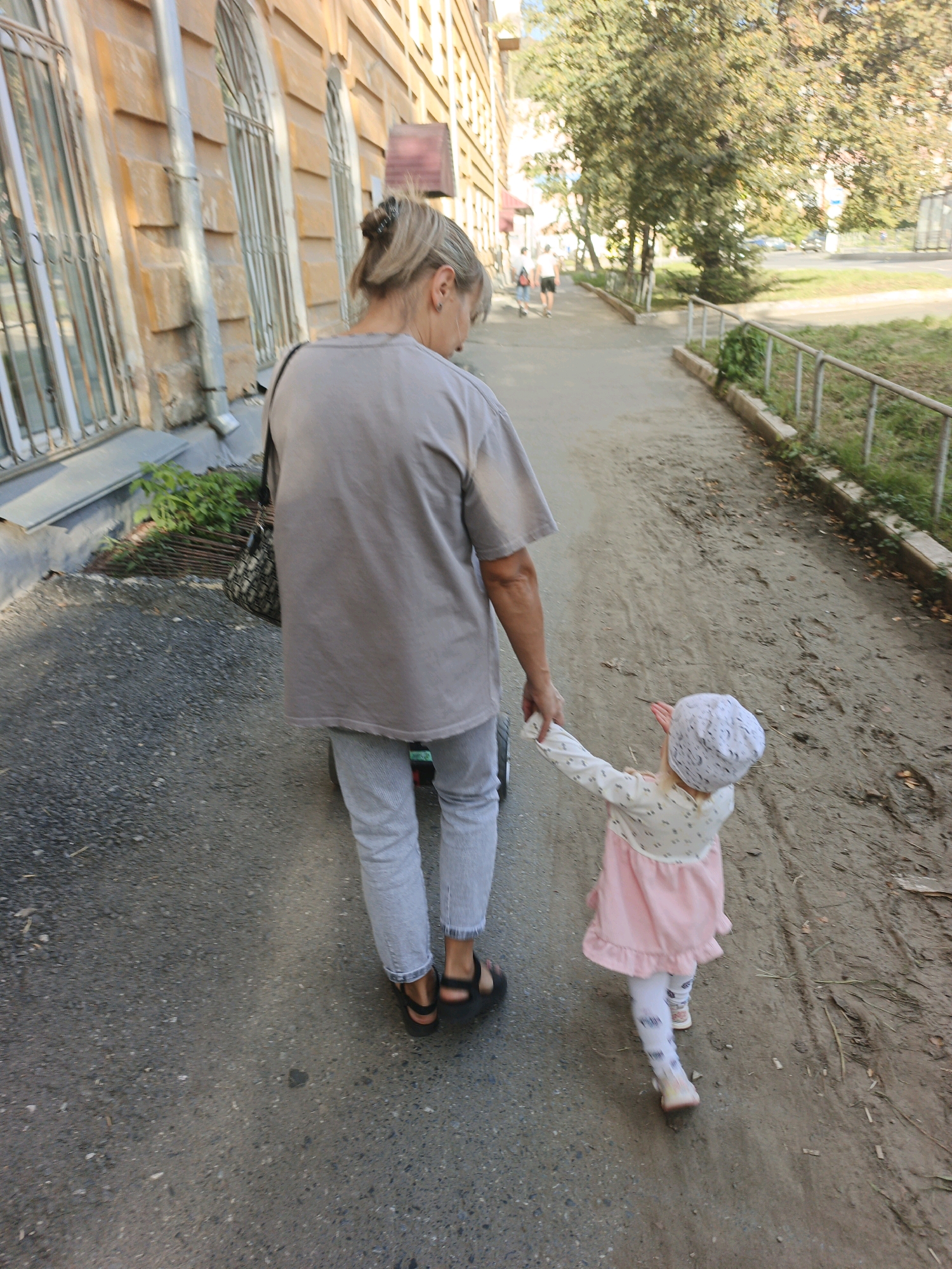 An adult, likely female based on hairstyle and clothing, is walking on a paved walkway with a young child. The adult is wearing a light-colored t-shirt and light-colored pants, and black sandals. They are holding the hand of the child, who is wearing a light-colored hat, a long-sleeved top with a pattern, and a pink skirt with white leggings. The adult appears to be pulling a small wheeled cart behind them. The walkway is bordered by a grassy area with a metal railing on one side and a building with arched windows and a textured facade on the other. Trees are visible in the background, suggesting an outdoor, urban setting. The scene is set in Zlatoust, Russia, specifically near the building that houses the Zlatoust local history museum and the Zlatoust State Museum of Art, as indicated by the architectural style of the building and the presence of trees and a walkway typical of such locations in Russian cities.