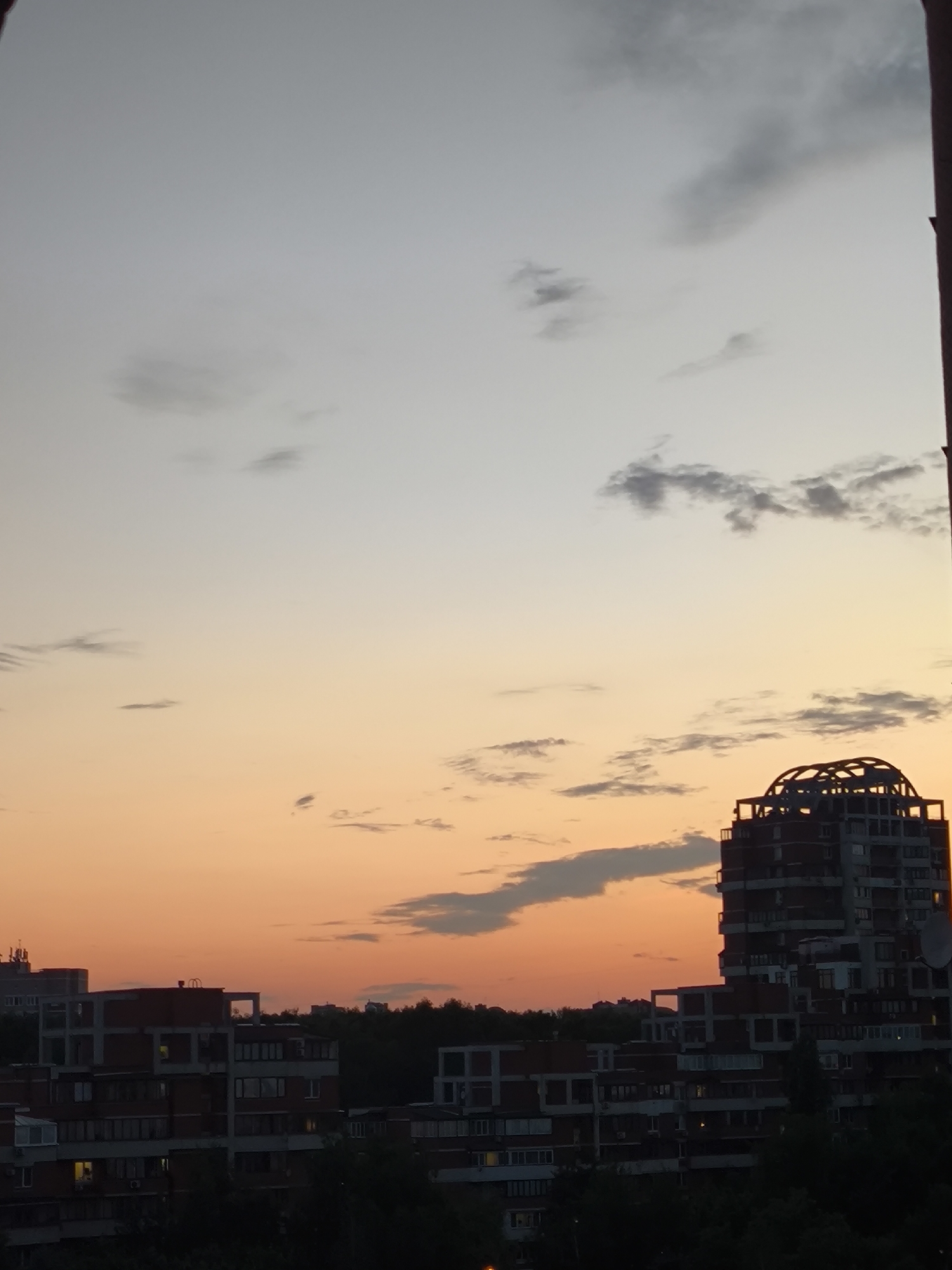 A cityscape during a sunset is depicted. The sky transitions from a pale grey at the top to a warm orange hue towards the horizon. Several scattered clouds are present. In the foreground, multiple apartment buildings are visible. One building on the right is taller than the others and features a distinctive circular, cage-like structure at its top. The buildings are made of brick and concrete, with visible windows and balconies. Trees line the base of the buildings, and the overall impression is one of an urban environment at dusk. The location is identified as Unknown, Unknown.