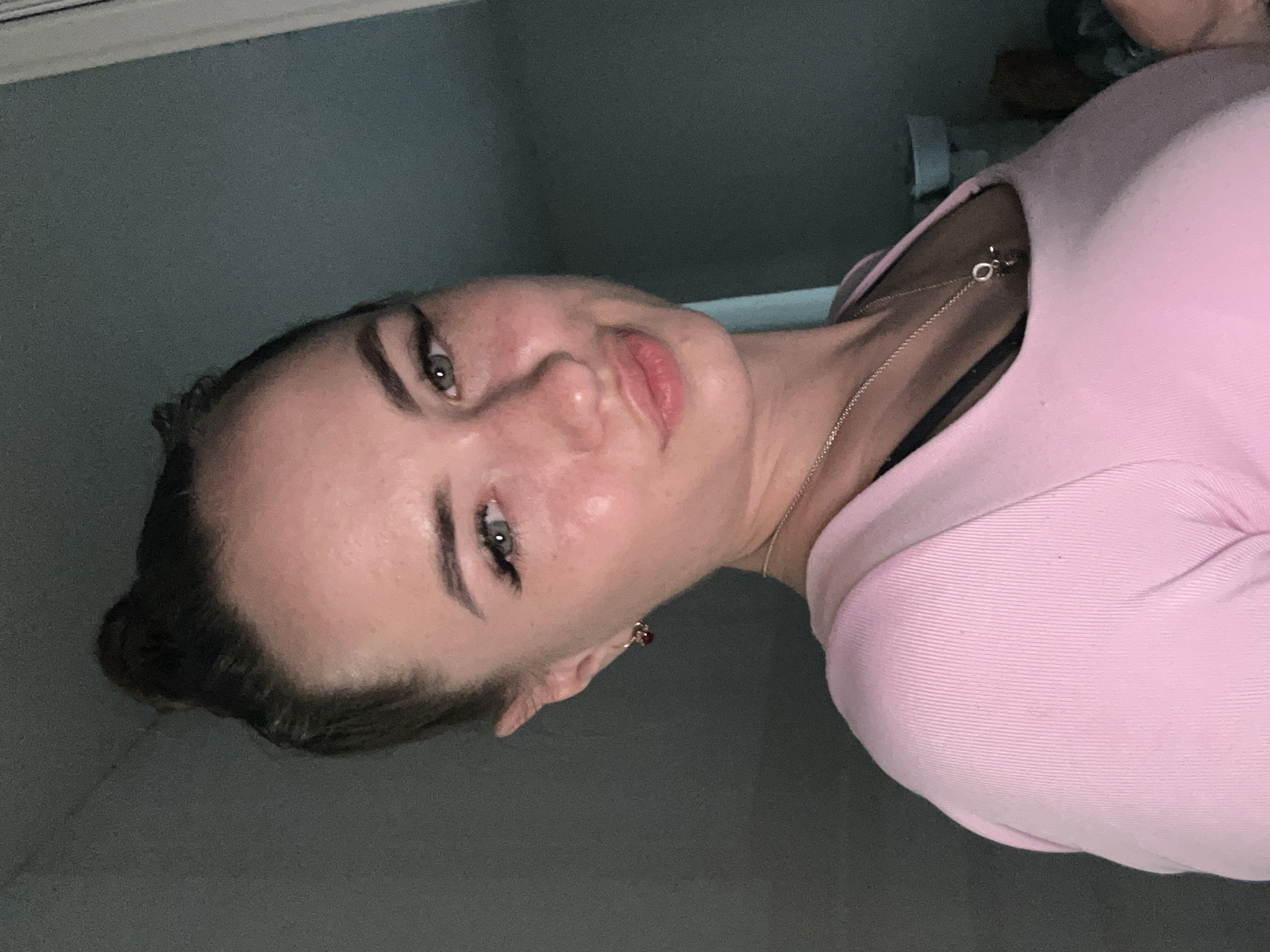 A young woman is pictured from the chest up, looking directly at the camera. She has her hair pulled up in a bun and is wearing a pink long-sleeved shirt. She has on makeup, including eyeliner and mascara. A necklace with a pendant is visible around her neck. The background appears to be indoors, with a wall behind her.