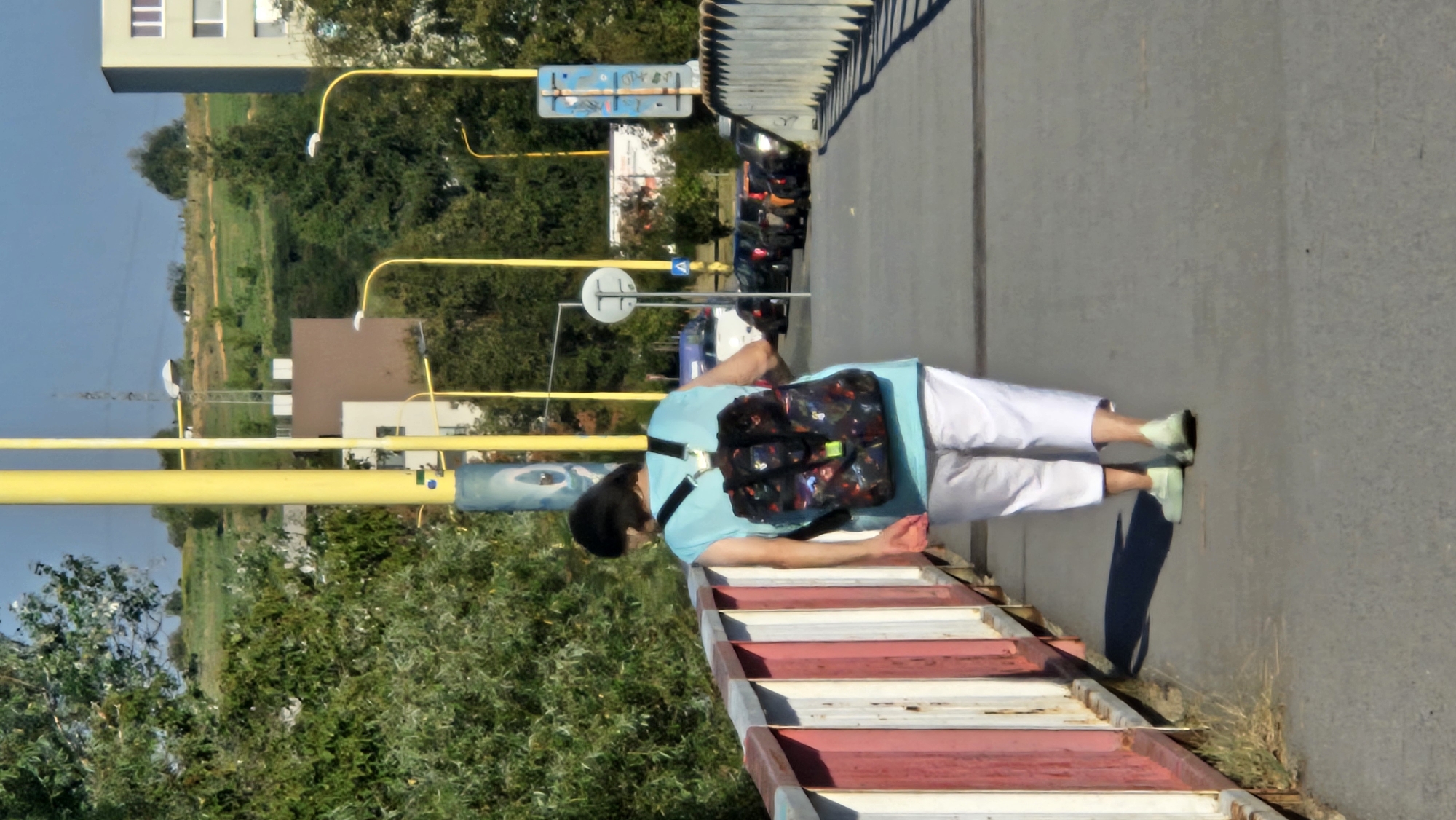 A woman walks across a pedestrian bridge in Prešov, Slovakia. She has short, dark hair and is wearing a light blue sleeveless shirt and white capri pants. A colorful backpack with a floral or abstract pattern is on her back. The bridge has a railing with vertical red and white panels on the left side. The walkway is paved with asphalt. In the background, there are trees, buildings, streetlights, and parked cars. The sky is blue with some clouds.