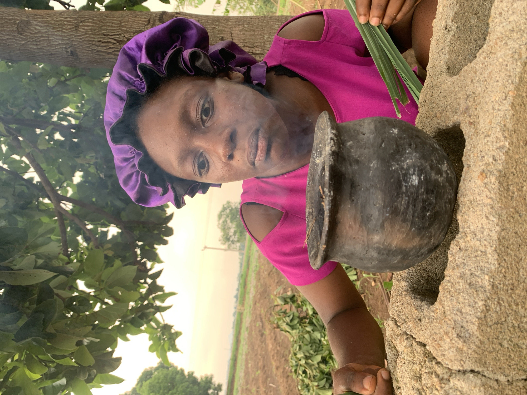 A woman wearing a purple head covering and a pink shirt stands outside in Agban, Nigeria. Smoke rises from a dark pot in front of her. She looks directly at the viewer. She holds long green leaves in her right hand. A rough stone structure is to her right. In the background is a field and trees.