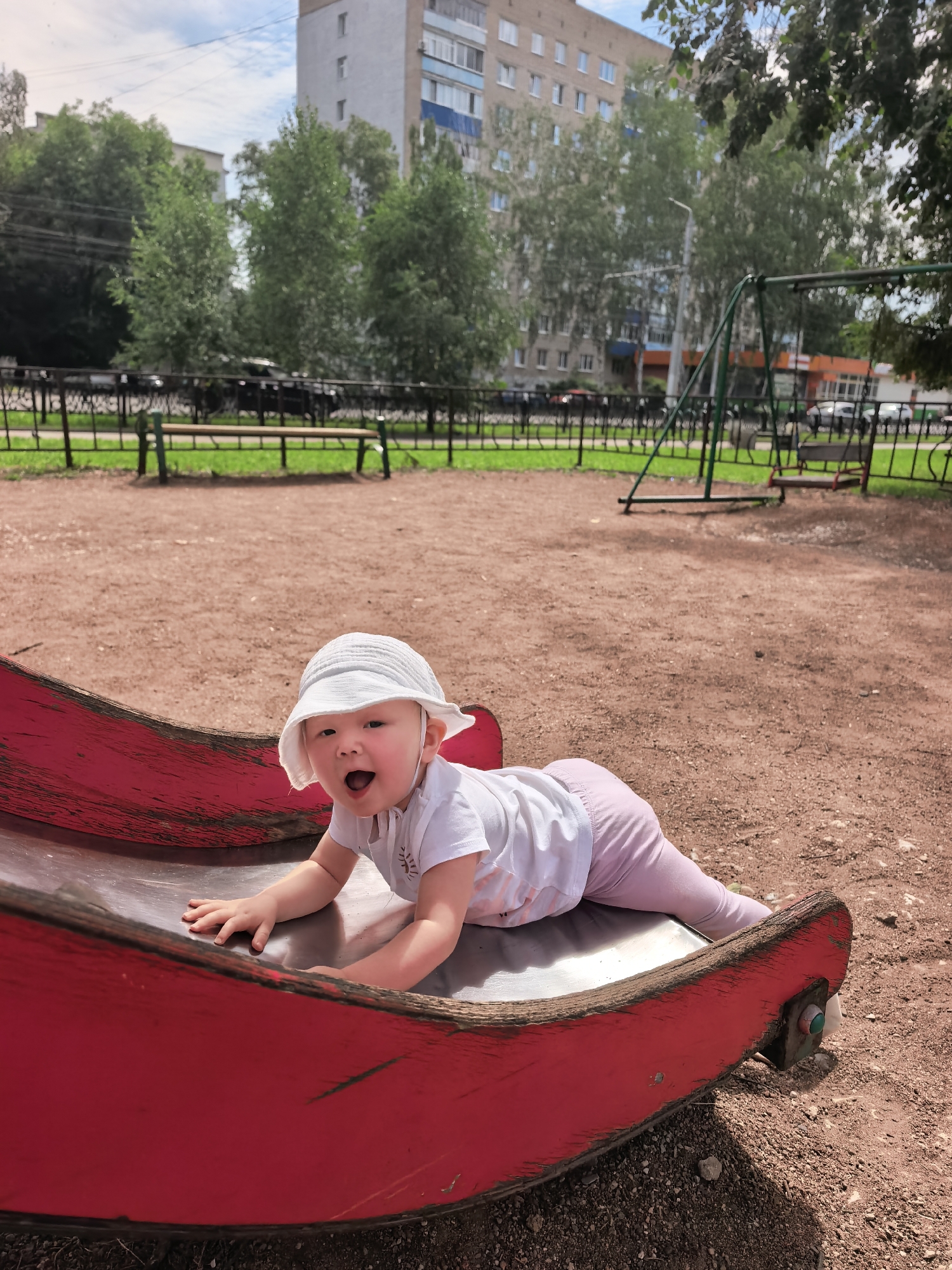 A baby wearing a white sun hat and a white t-shirt is on a red playground slide. The baby is on their stomach, reaching out with their hands. They are in a park with a swing set and a bench in the background. The park is located in the city of Sterlitamak, Russia. The sky is cloudy.