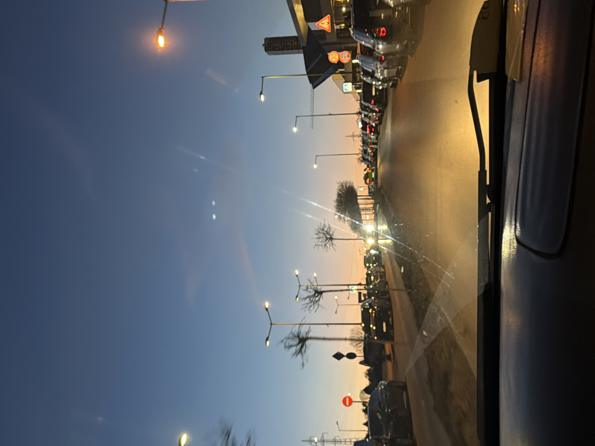 The image, likely taken from inside a vehicle, presents a view of a city street at dusk, potentially in Sofia, Bulgaria, given the presence of certain European-style signage and architecture. The sky transitions from a deep blue to an orange hue near the horizon. A line of stationary cars is visible along the right side of the road. Streetlights illuminate the scene, creating a contrast between the artificial light and the fading natural light. Signage, including road signs and possible business signs, is partially visible. Bare trees dot the roadside. The interior of the vehicle is partially visible on the right edge of the image, including the windshield and a portion of the dashboard.