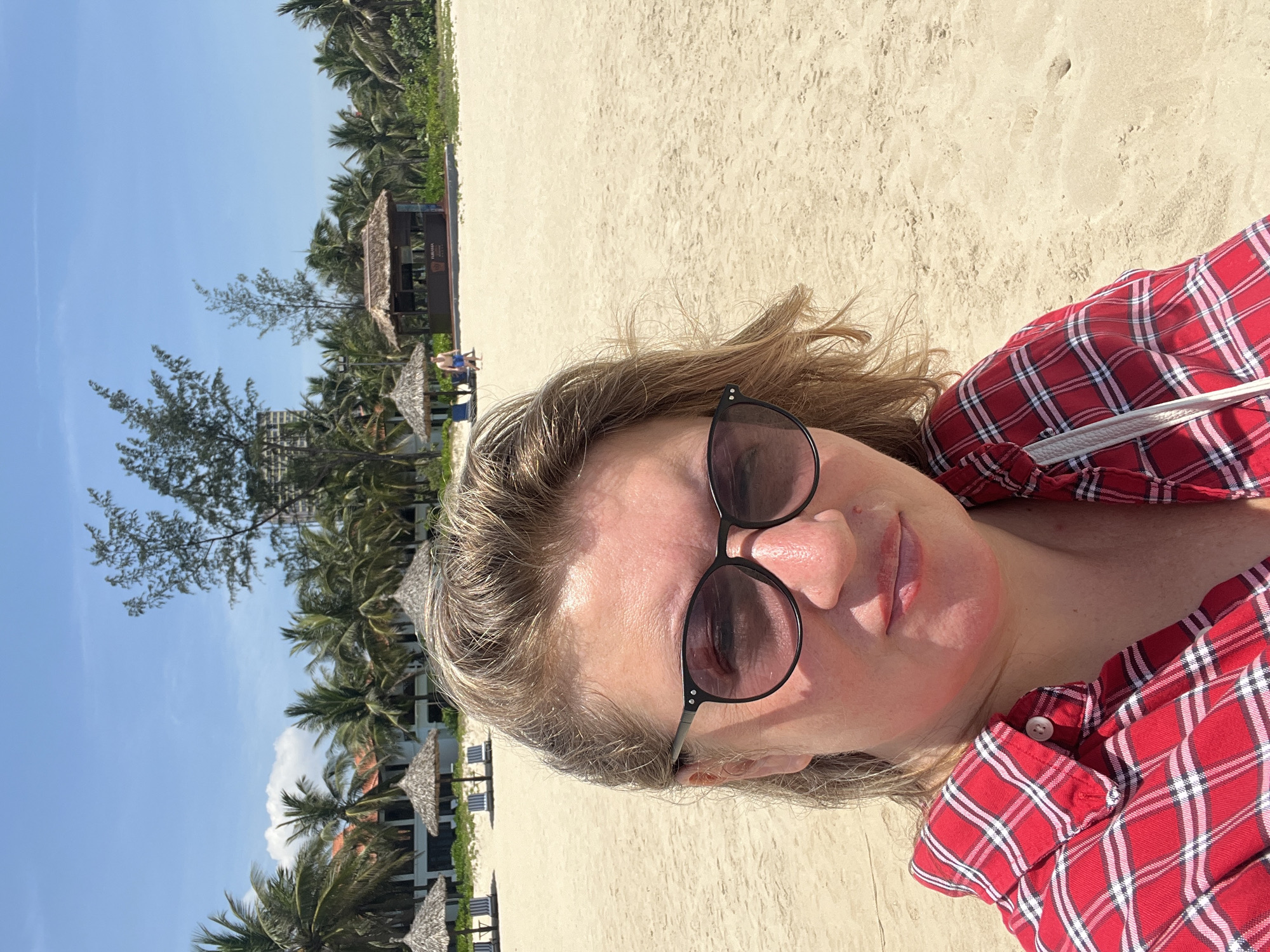 The image depicts a woman in a red plaid shirt and sunglasses lying on a beach. The beach is sandy, with some vegetation and palm trees visible in the background. A building with a thatched roof is also present. The location is likely a beach in Đà Nẵng, Vietnam.