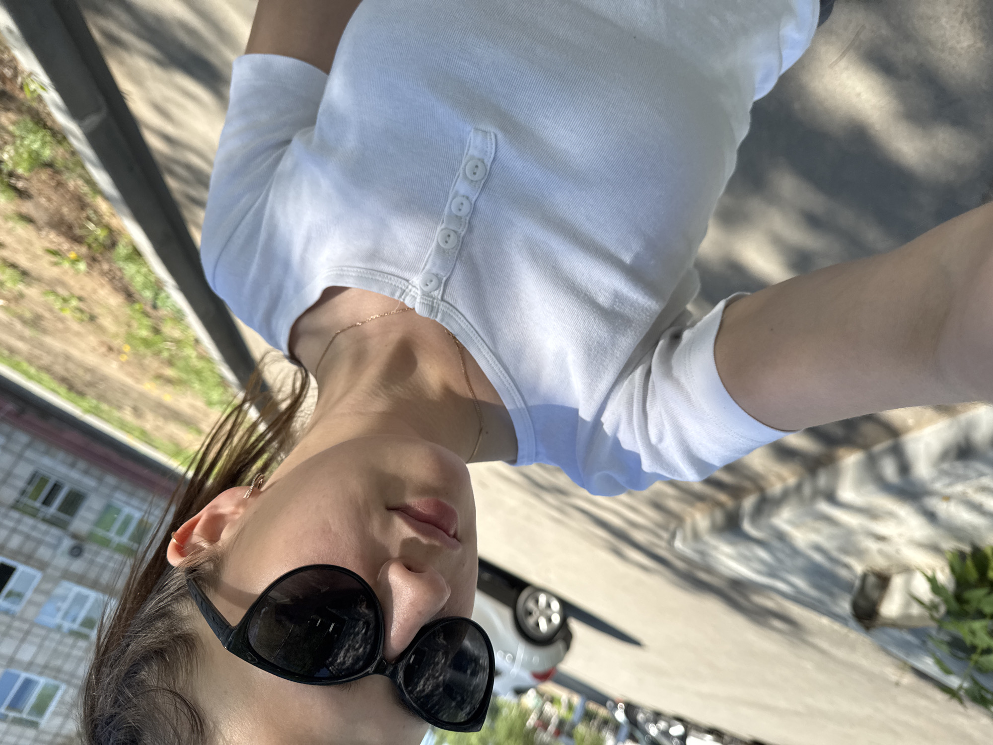 The image shows a selfie of a woman wearing sunglasses and a white button-down shirt. She is outside, likely in the city of Chaikovsky, Russia, judging by the style of apartment building visible in the background. The apartment building is a mid-rise structure, possibly built during the Soviet era, with white-framed windows. Behind the woman is a street with cars parked, and there are some trees and grass. The ground appears to be paved with what might be concrete or asphalt. The lighting suggests it's a sunny day.