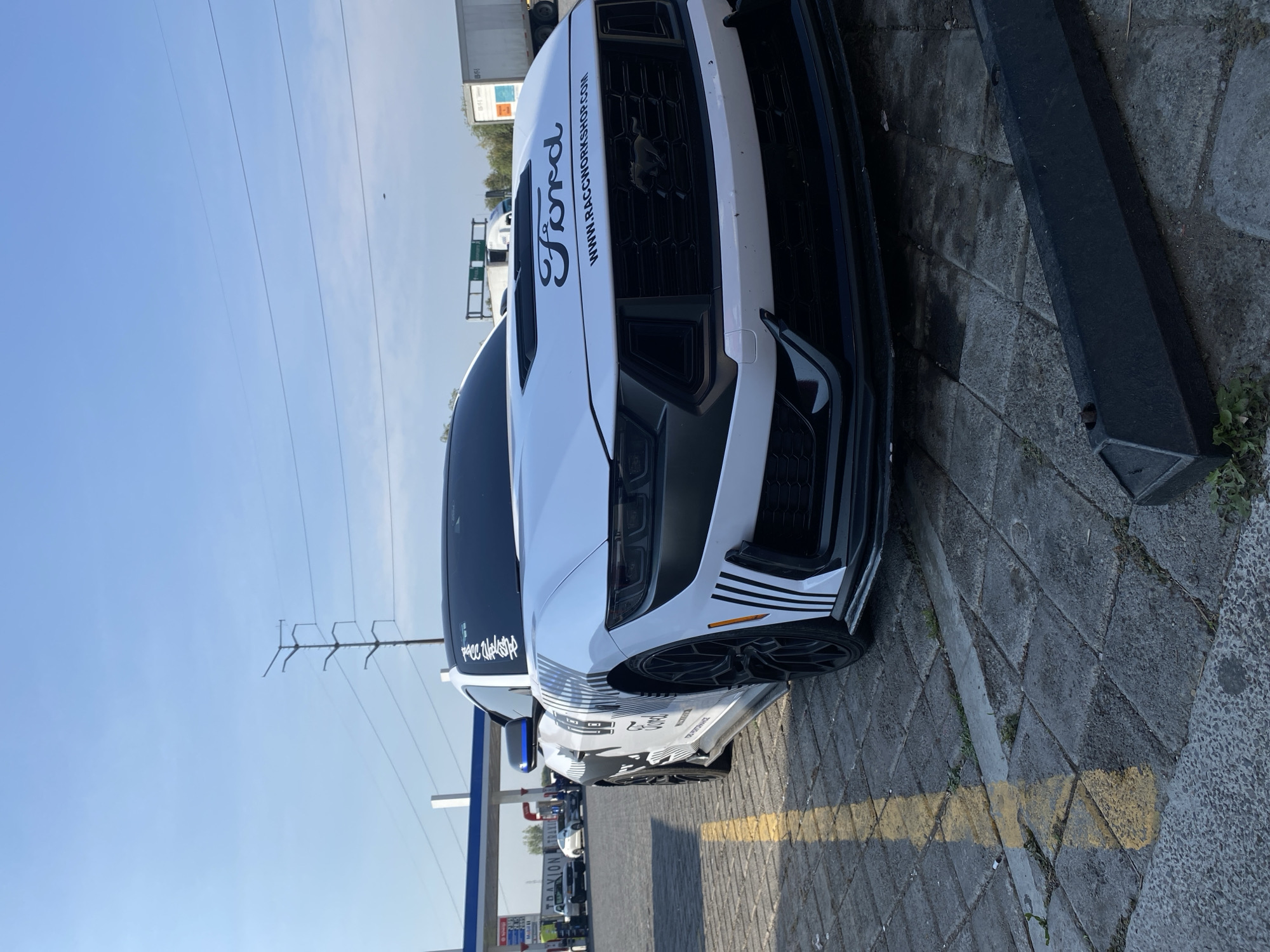 A white Ford Mustang with black decals is parked on a paved surface, likely a parking lot or roadside. The car has prominent "Ford" lettering on the hood and "www.raceworkshop.com" as well. The location appears to be at a gas station, identified by the gas pumps and pricing boards. The photo was taken during daylight hours with a clear, blue sky in the background. Power lines and utility poles are also visible in the background, indicating an urban or semi-urban setting. The presence of other parked cars suggests a public area. No people are visible in the immediate foreground. The exact location is Santa Ana Xalmimilulco, Mexico.