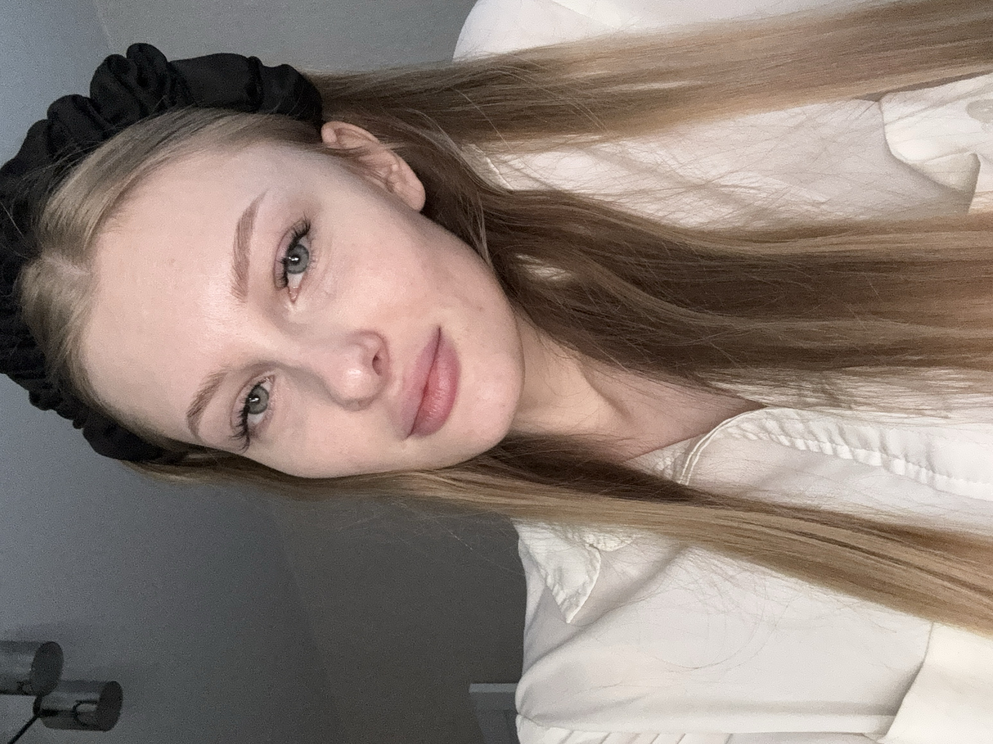 A young woman with long blonde hair, wearing a black headband and a white collared shirt, poses for a close-up selfie. Her face is turned slightly to the left, and she has a neutral expression. The background is a plain, neutral gray wall, and two light fixtures are visible in the upper left corner of the image. The lighting is soft and diffused, creating a subtle shadow on the left side of her face. The image appears to have been taken indoors, possibly in a residential setting. There are no specific landmarks or contextual clues that would indicate the location in the city of Perm, Russia.