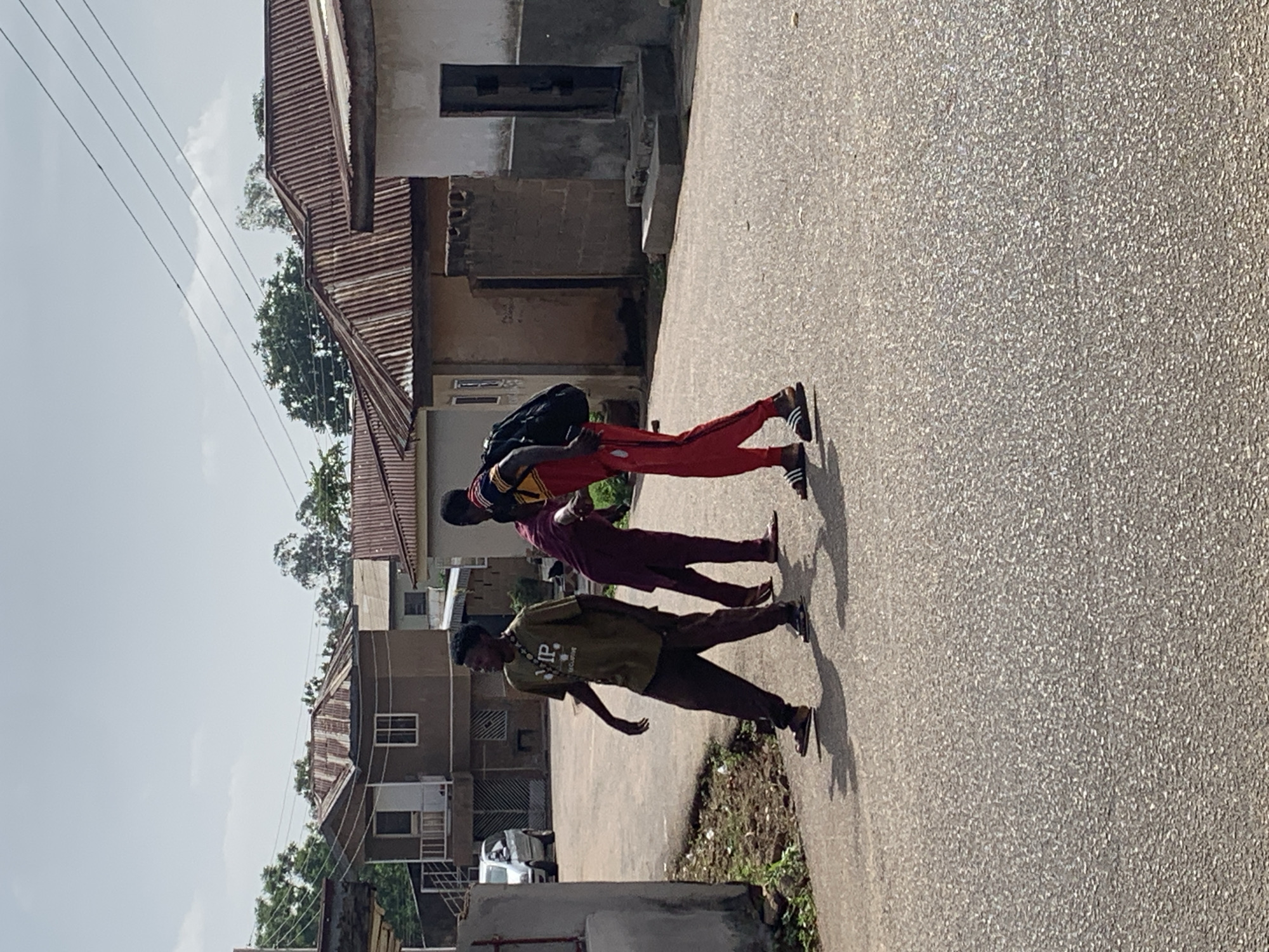 Three young men are depicted walking on a paved road in what appears to be a residential area in Jos, Nigeria. The image is taken outdoors on a sunny day, with clear to partly cloudy skies visible above and shadows cast by the subjects, suggesting late morning or early afternoon.
The leftmost man, in the foreground, wears a dark olive-green t-shirt with "VIP" printed prominently on the chest, along with dark pants and dark shoes. He is looking slightly downwards. The man in the middle wears a maroon or dark red tunic-style shirt and dark pants, also with dark shoes, and is looking straight ahead. The third man, towards the background, is dressed in a red tracksuit with black stripes, a multi-colored shirt (yellow, black, red), and black and white striped Adidas sneakers. He carries a black backpack. All three men have dark complexions and short hair.
The road is grey asphalt, textured with small aggregate. On the left side of the road, there's a strip of dry earth and sparse green vegetation. The background features several single and two-story buildings with corrugated metal roofs, some showing signs of rust. The buildings are constructed with light-colored concrete or stucco. Overhead, utility lines span horizontally across the sky. The overall scene depicts a casual daily activity in an urban or semi-urban setting.