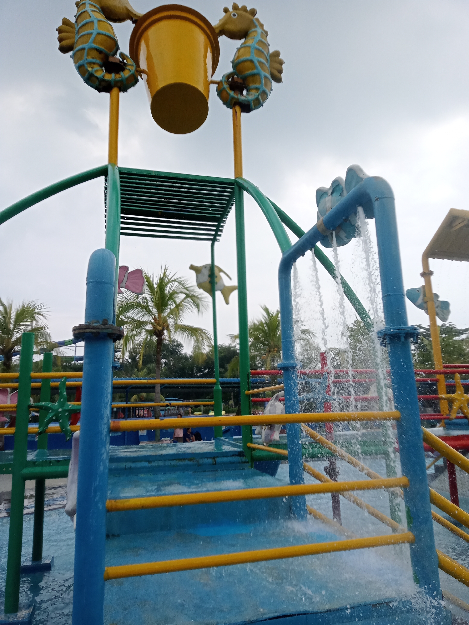 The image depicts a water park attraction in Baru, Indonesia. The main subject is a large, yellow bucket suspended by two seahorse figures, which is part of a water play structure. Water is actively cascading from pipes, creating a spray effect. The structure is painted in bright colors, primarily blue and yellow, with green supporting poles. Palm trees and lush greenery are visible in the background, suggesting a tropical or subtropical climate. The sky is overcast, indicating a cloudy day. Several people are present in the background, appearing to be enjoying the water park facilities. The scene captures a moment of play and recreation within a water park environment.