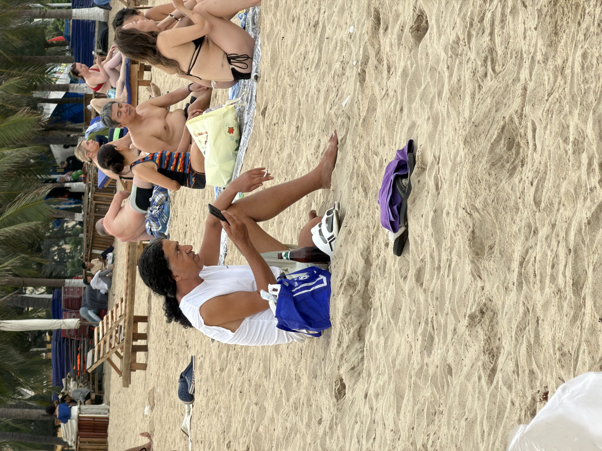 The image depicts a beach scene in Nha Trang, Vietnam. In the foreground, a man with dark, curly hair wearing a white sleeveless shirt and light-colored shorts sits on the sand, looking at his phone. Next to him is a blue tote bag and a brown bottle. Further in the background, several other individuals are seated on the sand, some on loungers. They appear to be in casual beach attire. Palm trees are visible in the background, indicating a tropical or subtropical location. The sand is light brown and rippled.
