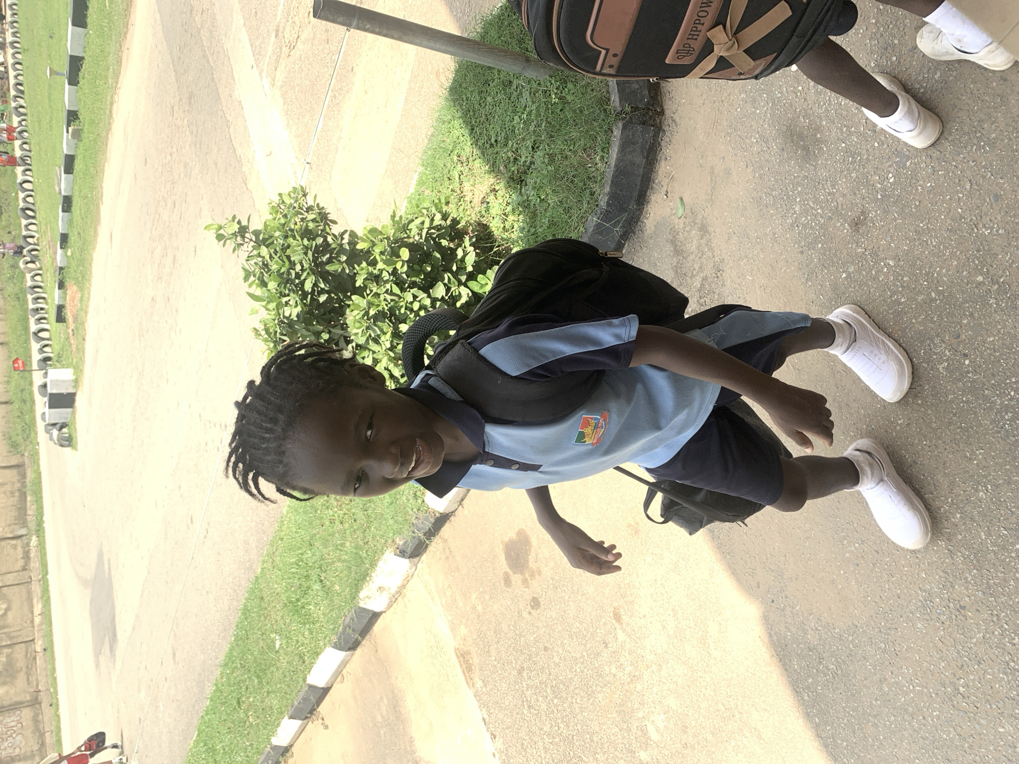 A young boy, likely of African descent, stands on a paved surface, possibly a sidewalk, and is looking towards the camera with a smile. He is wearing a light blue polo shirt with dark blue trim and navy blue shorts, along with white socks and white sneakers. He has a black backpack on his shoulders and is holding a black bag in his left hand. The background shows a grassy area and a road, with a line of tires and concrete barriers along the side of the road. The lighting suggests it is daytime and sunny. The overall scene appears to be an outdoor public space, possibly a school or recreational area, with other children and adults faintly visible in the distance on the road. The environment appears to be a city, potentially Abuja, Nigeria, given the context of the prompt.