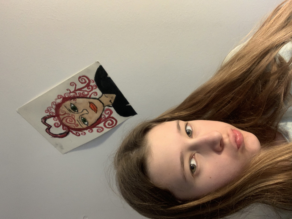 The scene depicts a young female indoors within the Vozdovac Urban Municipality, Serbia. The female, of apparent adolescent age, has long, straight, light brown hair with a central parting and light-colored eyes. She is positioned facing the camera, with her head slightly tilted to her right and her lips formed into a pout. She wears a light-colored, possibly light blue or grey, top. Situated on the wall behind and above her, offset to the right, is a rectangular canvas painting. The artwork portrays a stylized female face with bright red, curly hair, including a prominent heart shape formed at the top of the hairstyle. The painted figure has green eyes and orange lips, and is depicted wearing a black top against a white background. The painting is mounted askew on a plain, light-colored wall. The image appears to be a self-portrait taken in an interior environment.