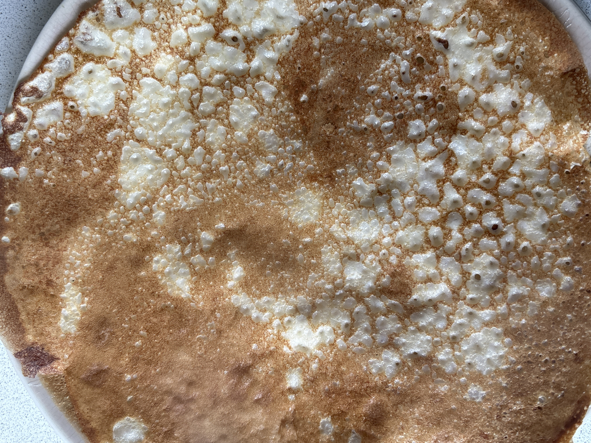 A close-up, overhead shot of a cooked pancake or crepe resting on a white plate. The pancake is mostly circular in shape and golden brown with numerous white spots. The surface of the pancake has a blistered texture. The plate has a slightly raised rim. The background is a light gray surface. There is no indication of the location of the image.