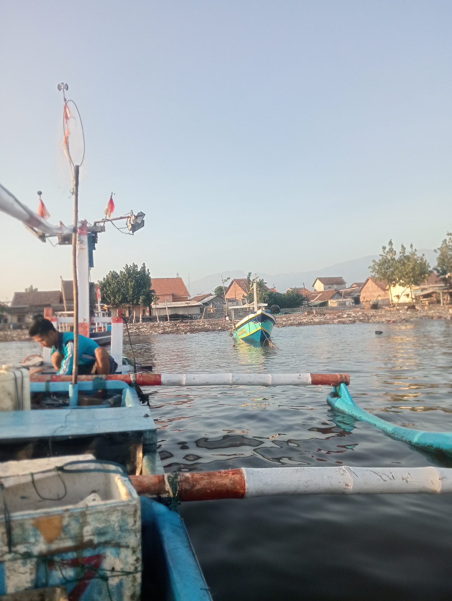In Besuki, Indonesia, on the morning of an unspecified date, a fishing boat is moored near a coastline littered with debris. The sky is clear and pale blue. A young man, wearing a blue shirt and black shorts, crouches on the deck of the boat, engaged in an unknown task. His posture suggests he is focused on something on the boat's floor.
The boat itself is partially visible, with its blue hull and wooden structure. Part of a white bucket with red and blue markings is in the foreground, suggesting it is used for storing fishing gear or catch. Another fishing boat, painted blue and yellow, floats in the water in the middle ground, appearing empty.
The shoreline is densely populated with houses and trees. A large amount of trash is visible along the waterline and on the beach, indicating environmental pollution. In the background, a mountain range is faintly discernible against the horizon. The scene is captured from the perspective of someone on another boat, looking towards the shore. The presence of a mast with flags on the visible boat suggests it is prepared for or has just completed a fishing trip. The overall impression is one of a working fishing community facing environmental challenges.