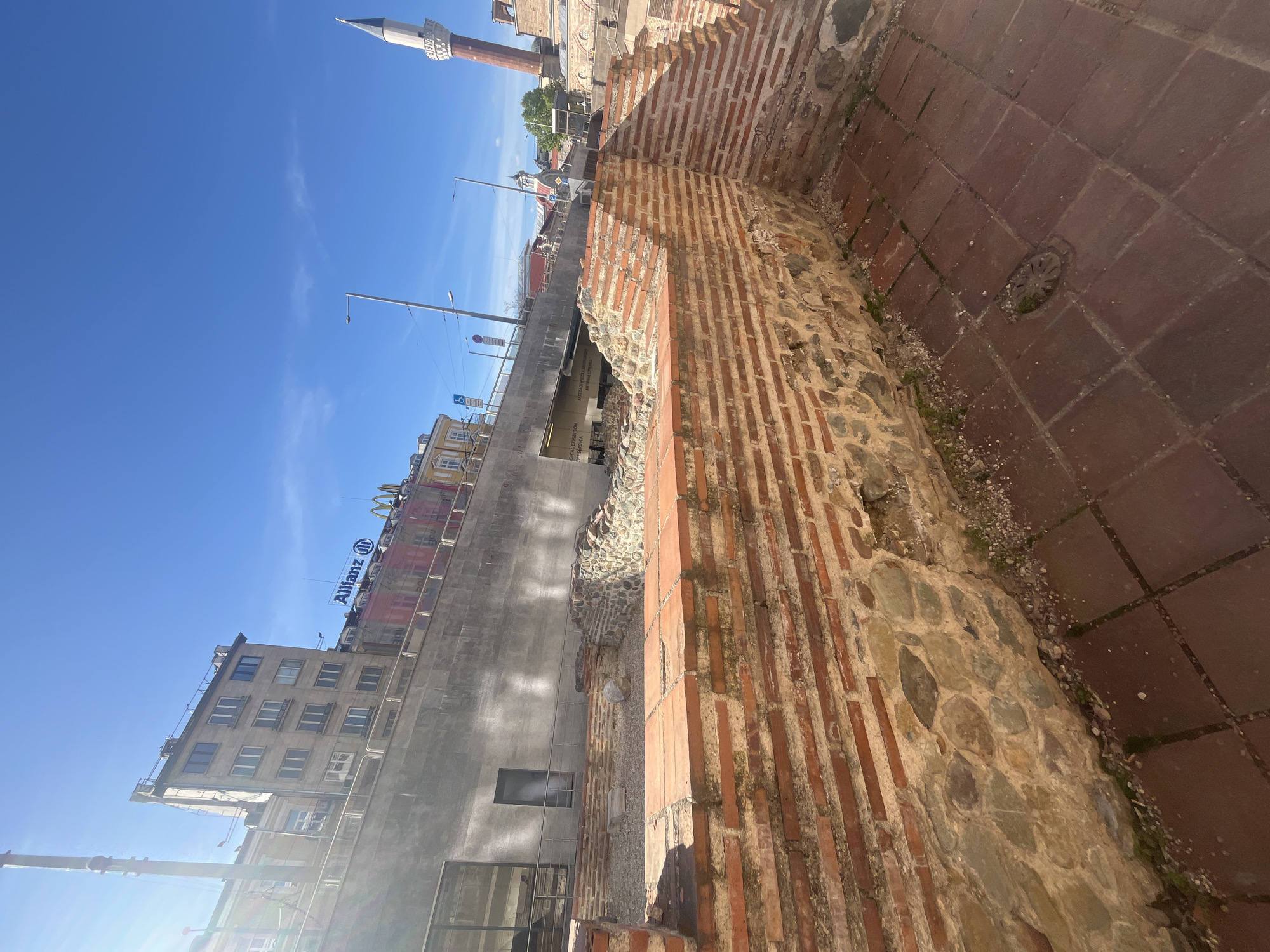 This image shows an outdoor scene in Sofia, Bulgaria. The foreground features a brick and stone wall, likely part of an archaeological site. The ground is paved with large, reddish-brown tiles. In the background, modern buildings are visible, including one with signs for "Allianz" and a McDonald's logo. Further back, a minaret is seen against a blue sky. The scene appears to be captured in daylight under sunny conditions.