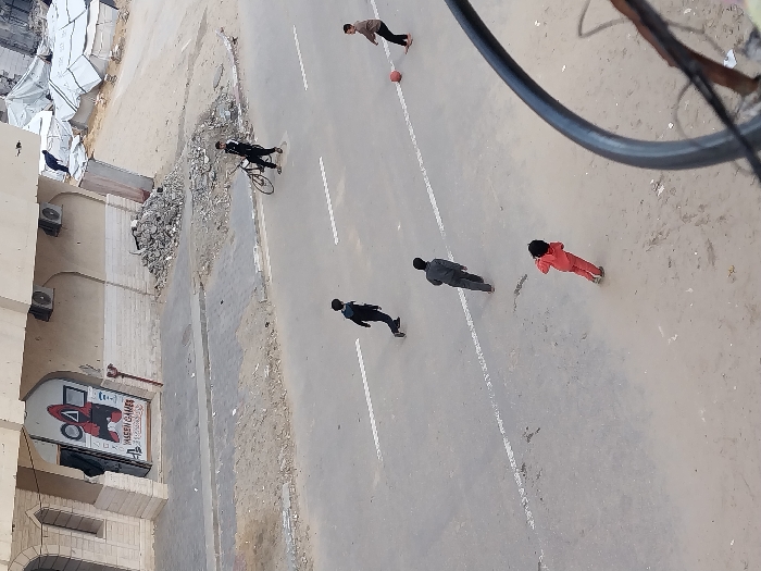 The image, captured from a high vantage point, depicts a street scene in Khan Yunis, Palestinian Territory. In the immediate foreground, at the bottom right, a portion of a dark, curved metal object, likely an antenna frame, is visible. The central area features a paved asphalt road, marked with two worn, broken white lines running horizontally. Five individuals are present on the road: one individual, appearing to be an adult or teenager, in dark clothing, stands on the right side holding a black bicycle. Four younger individuals (children or adolescents) are distributed across the road. One child in black clothing moves leftward, followed by another in grey clothing also moving leftward. A third child in red clothing stands stationary near the bottom-left. On the far right, a fourth child in brown/tan clothing is positioned near a red ball on the road. The children appear engaged in a game involving the ball.
In the background on the left, a light-colored, multi-story building with arched architectural details is visible. Its ground-floor entrance is decorated with a "Squid Game" theme featuring a circle, triangle, and square, and includes text indicating "YASEEN GAMES". Two external air conditioning units are mounted on the building's facade. A paved sidewalk runs along the front of this building, separated from the street by a dirt strip containing a pile of construction debris. To the right of the building and beyond the street, the background transitions to an area of disturbed earth and scattered rubble. Further into the distance on the right, several temporary structures, resembling tents or makeshift shelters made of white tarpaulin, are visible, with a dark-clad figure standing atop one of these structures or adjacent debris.