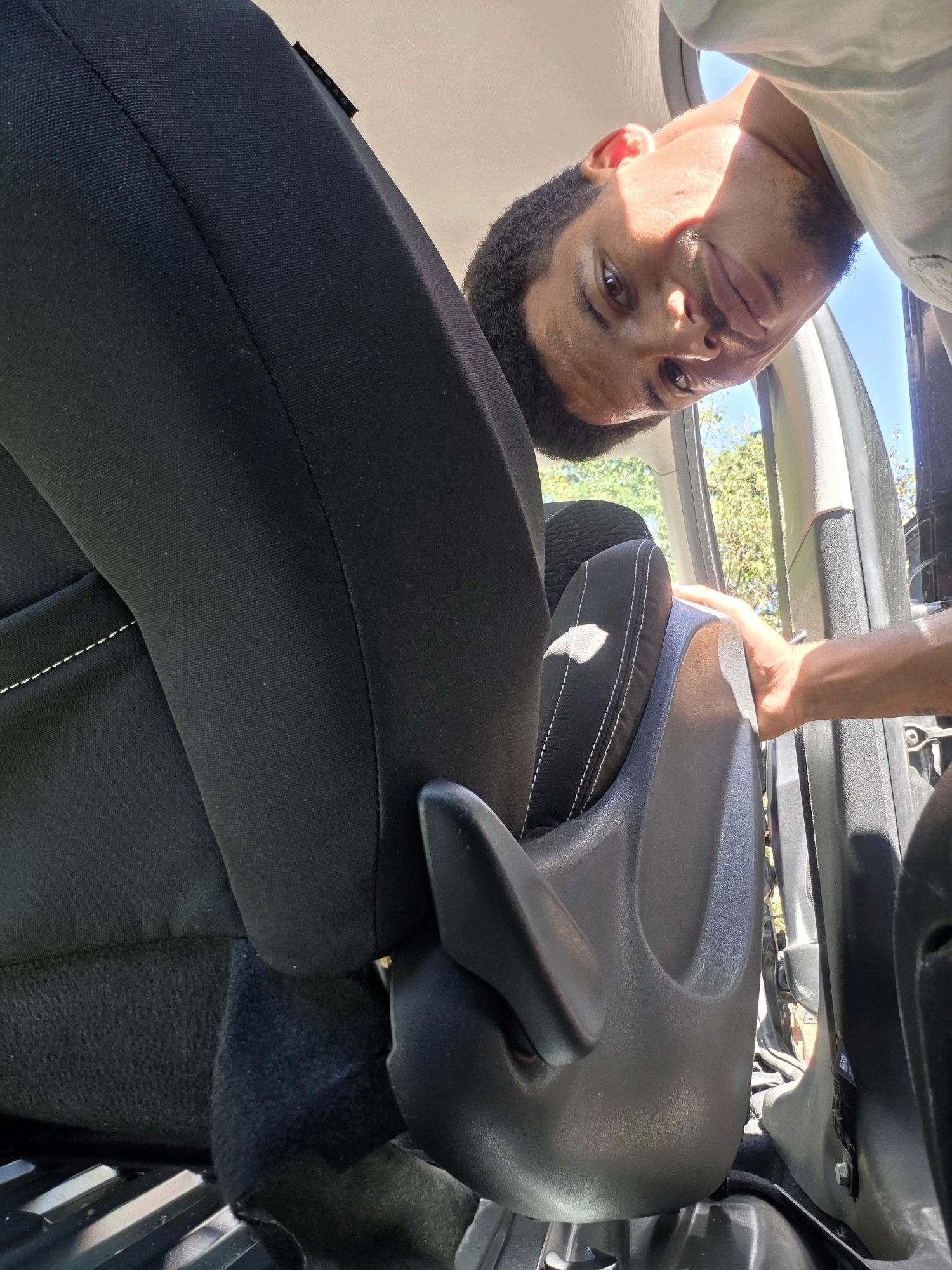 A young man leans into the interior of a car, looking upwards at the camera. He has short, dark, curly hair and is wearing a light-colored t-shirt. His face is partially obscured by shadows and the angle of his head. The car's interior is visible, including black fabric seats with white stitching and a dark gray plastic trim piece. Sunlight streams in from the open car door, illuminating the scene. The background outside the car shows green foliage and a clear blue sky. The scene is set in Varna, Bulgaria.