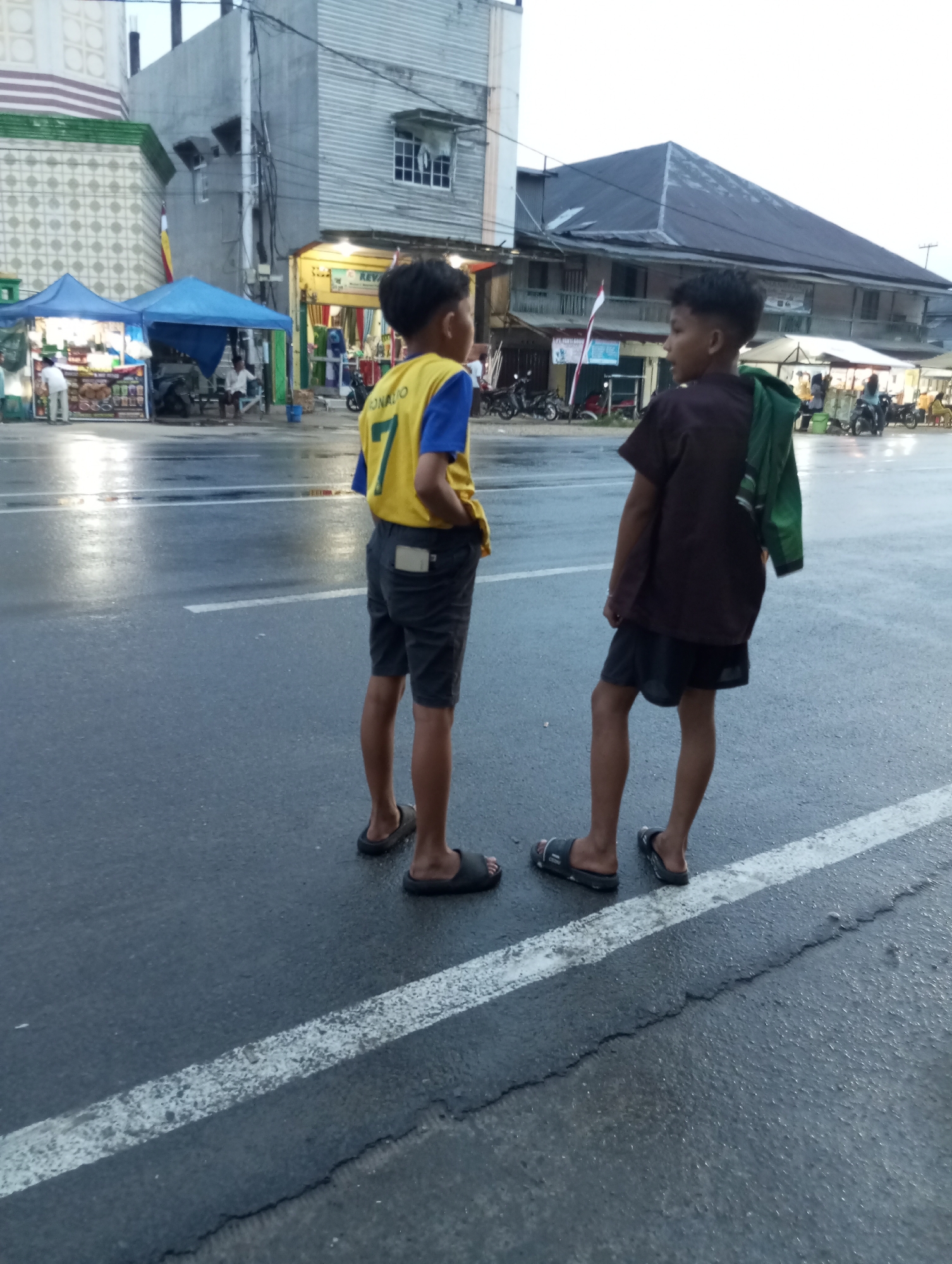 Two young boys stand on a wet asphalt road in Air Molek, Indonesia. The boy on the left wears a yellow and blue jersey with the number "7" and the name "CRONALDO" visible. He also wears dark shorts. The boy on the right wears a dark t-shirt and dark shorts, with a green garment slung over his shoulder. They are facing each other, appearing to be engaged in conversation. In the background, there are buildings, tents, and motorcycles, suggesting a market or roadside area. The sky is overcast, indicating a cloudy day.
