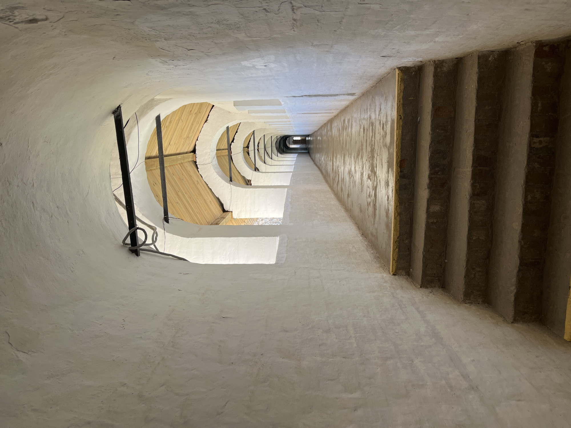 The image depicts a long, arched corridor with white-plastered walls and a wooden roof structure visible through the arches. The corridor floor is paved and appears wet or reflective, suggesting it might have recently rained or been cleaned. In the foreground, there are several stone steps leading up into the corridor, with yellow markings on the edges of the steps. The corridor recedes into the distance, with a series of repeating arches creating a sense of depth and perspective. The lighting suggests it is daytime, with natural light entering from the openings of the arches and potentially from the end of the corridor. There are no people visible in the image. The environment appears to be an outdoor or semi-outdoor passageway, possibly part of a historical building or complex, given the architectural style. The location is identified as Novo-Nikolskoye, Russia.