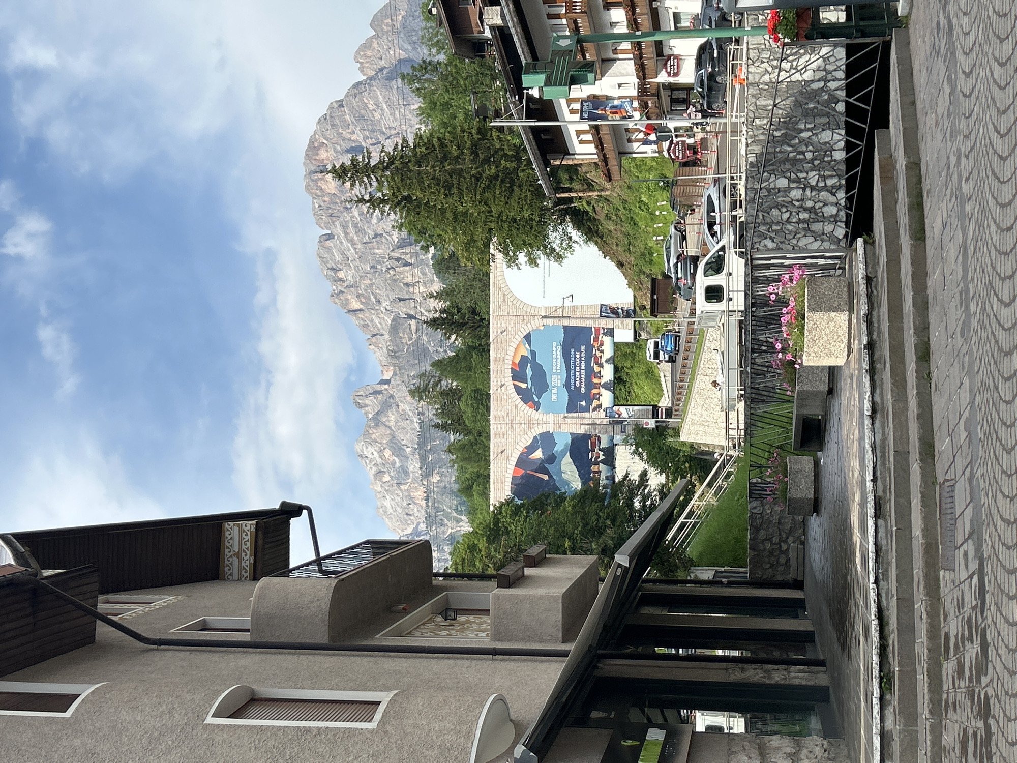 This media file captures a vertical outdoor scene showcasing a charming mountain town with a majestic mountain range in the background. The town features traditional architecture with wooden balconies and pitched roofs, nestled amidst lush green trees. In the foreground, a textured paved walkway leads up a set of stairs, with a stone wall and decorative planters adding to the aesthetic.
Dominating the middle ground is a large archway adorned with vibrant billboards. One billboard prominently displays text in Italian, including "CORTINA 2026 GIOCHI OLIMPICI PARALIMPICI" which indicates a connection to the Cortina 2026 Olympic and Paralympic Games. The billboards also feature artistic imagery with a blend of abstract shapes and silhouettes of people, suggesting a celebration or promotion related to the event.
The weather appears to be pleasant, with a partly cloudy blue sky suggesting it's daytime. The overall atmosphere is serene and picturesque, conveying a sense of community pride and anticipation for the upcoming games. The scene is set in what appears to be a European mountain resort town, with the imposing mountain range serving as a dramatic backdrop. No people are clearly visible in the image, but the presence of parked cars and the general setup of the town suggest it's a populated area.
