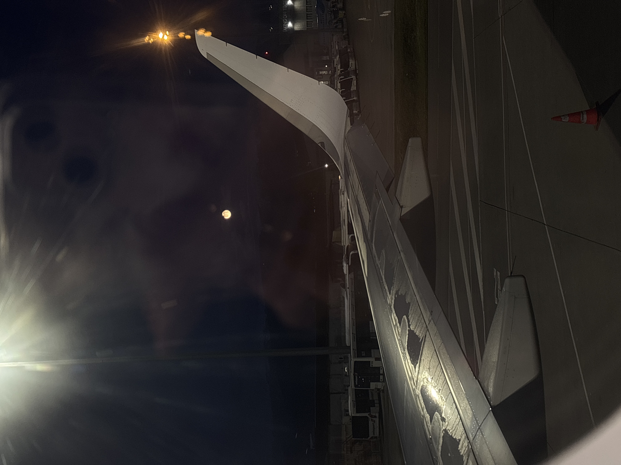 The image depicts the wing of an airplane at night, viewed through the plane's window. Outside, a few lights are visible in the distance, suggesting an airport setting. The ground shows tarmac markings and an orange traffic cone is visible on the ground outside the window.