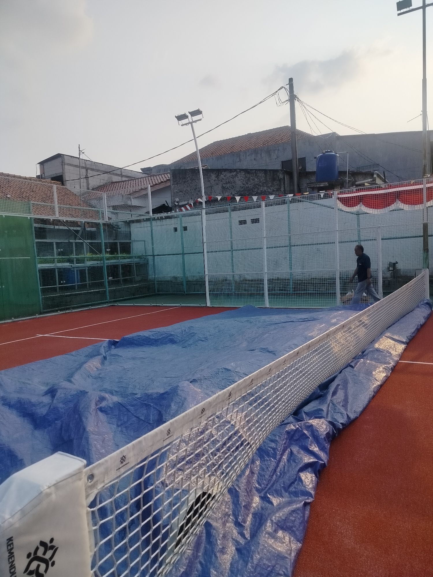 A padel tennis court in East Jakarta, Indonesia, is partially covered with a large blue tarp. A white net spans the width of the court, with the tarp draped over it and extending across the playing surface. A man, wearing a dark shirt and light-colored pants, is walking away from the net and towards the back of the court. He appears to be heading towards a fence enclosure that surrounds the court, which is also bordered by buildings and a water tank. The sky is overcast.
