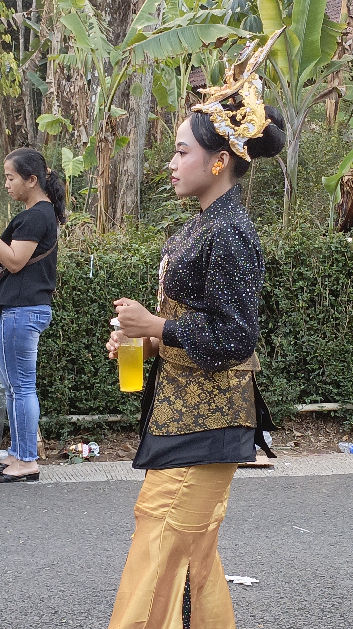 In Krajan Lor, Indonesia, a young woman with a golden headdress adorned with intricate designs stands in profile, facing left. She wears a black, sparkly blouse and a gold brocade sash. A bright yellow drink in a plastic cup is held in her right hand. Behind her, another woman in a black t-shirt and blue jeans is partially visible, looking down. The backdrop consists of lush green foliage and trees, suggesting an outdoor setting.