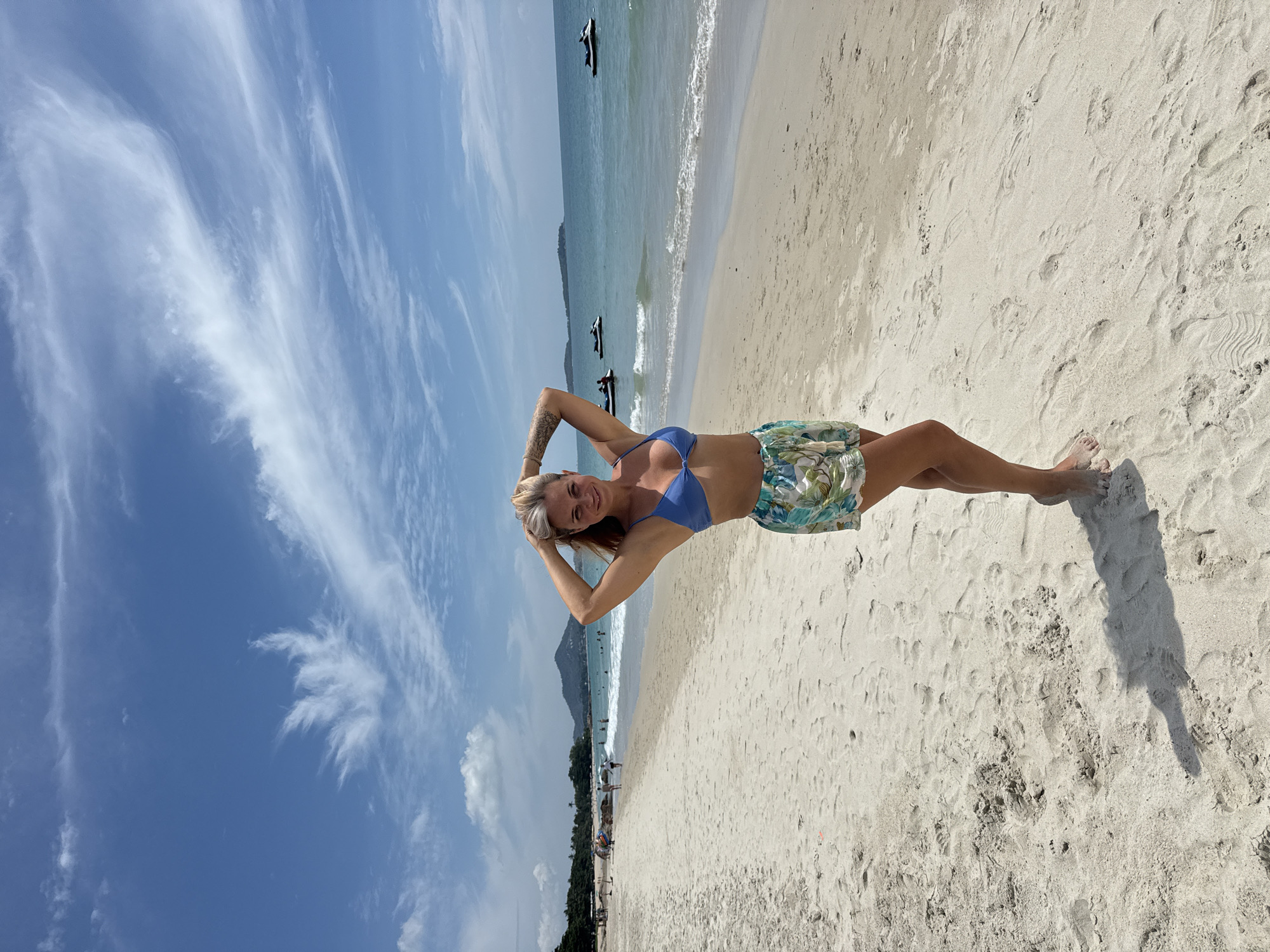 A young woman with blonde hair, wearing a blue bikini top and floral shorts, stands on a sandy beach in the Ko Samui District of Thailand. She has her hands on her head and is smiling at the camera. The beach is wide and sandy, with gentle waves lapping at the shore. In the background, there are several jet skis in the water and palm trees lining the coast. The sky is blue with scattered white clouds.